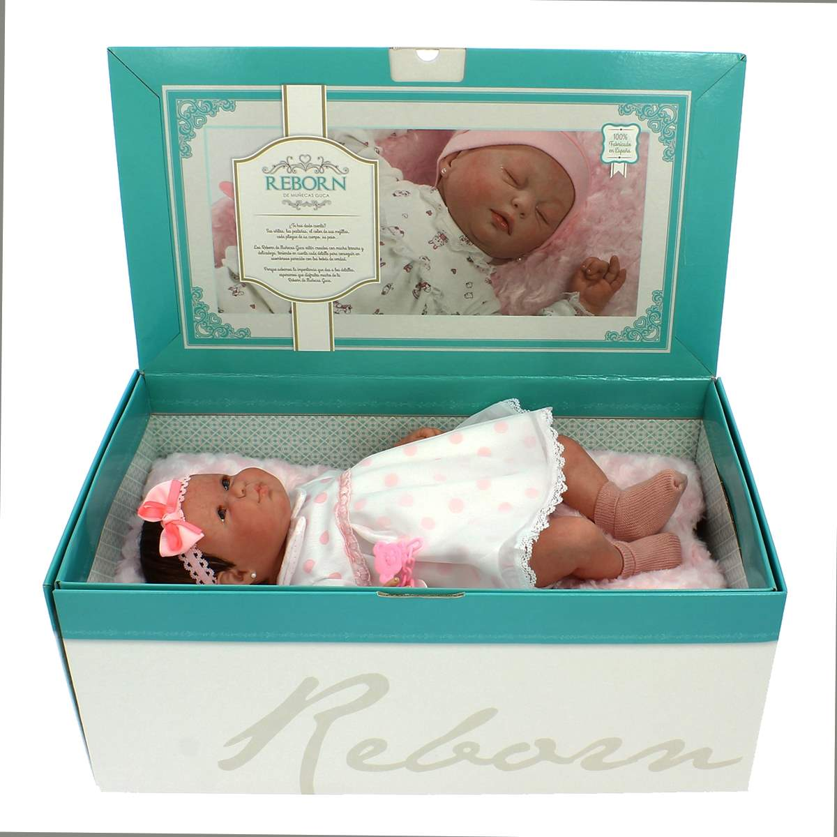 Una muñeca bebé en una caja. Viste un vestido blanco con lunares rosas y un lazo rosa. La caja dice "Reborn". La parte superior muestra un bebé durmiendo.
