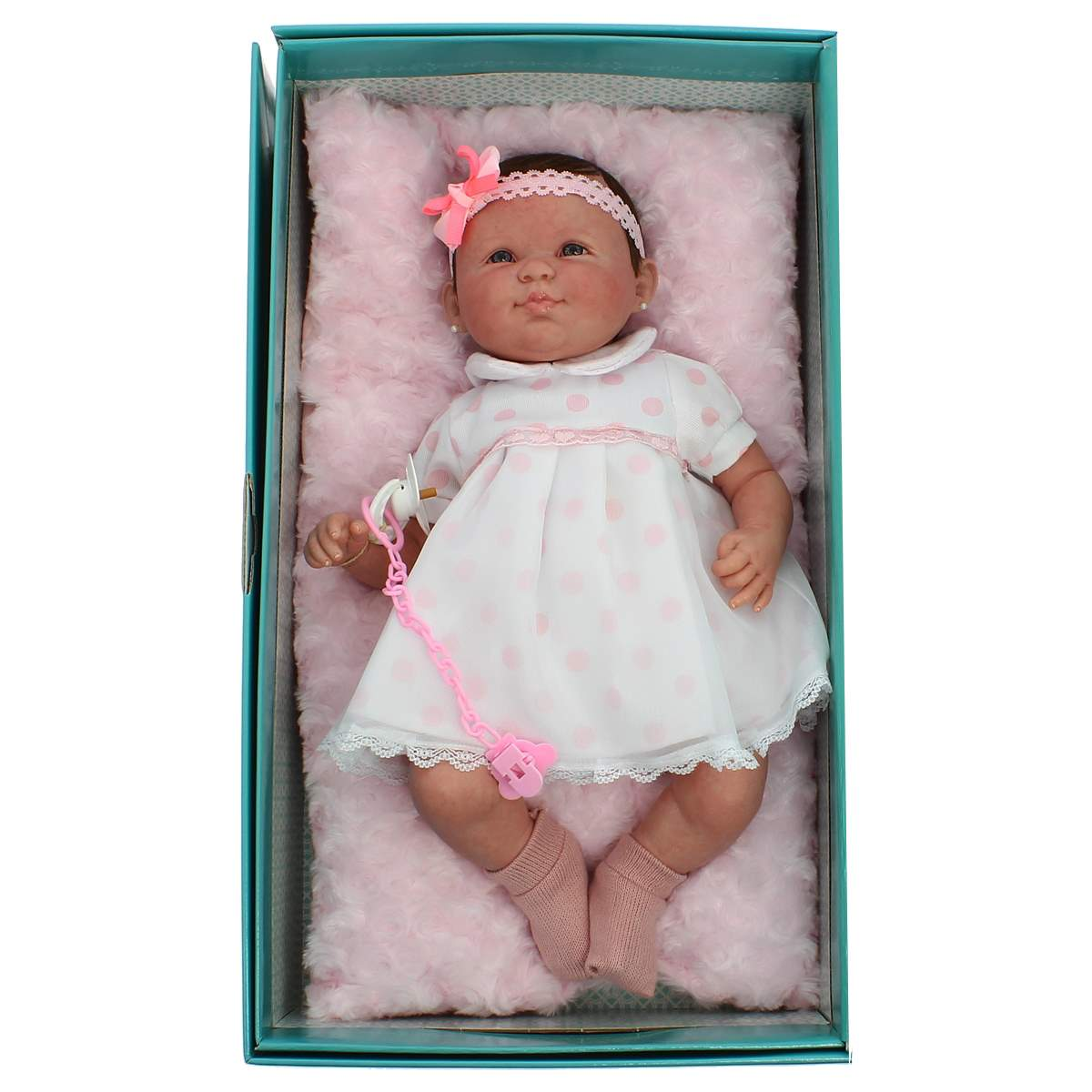 Una muñeca bebé en una caja, con un vestido blanco con lunares rosas, calcetines rosas, una diadema y un chupete.