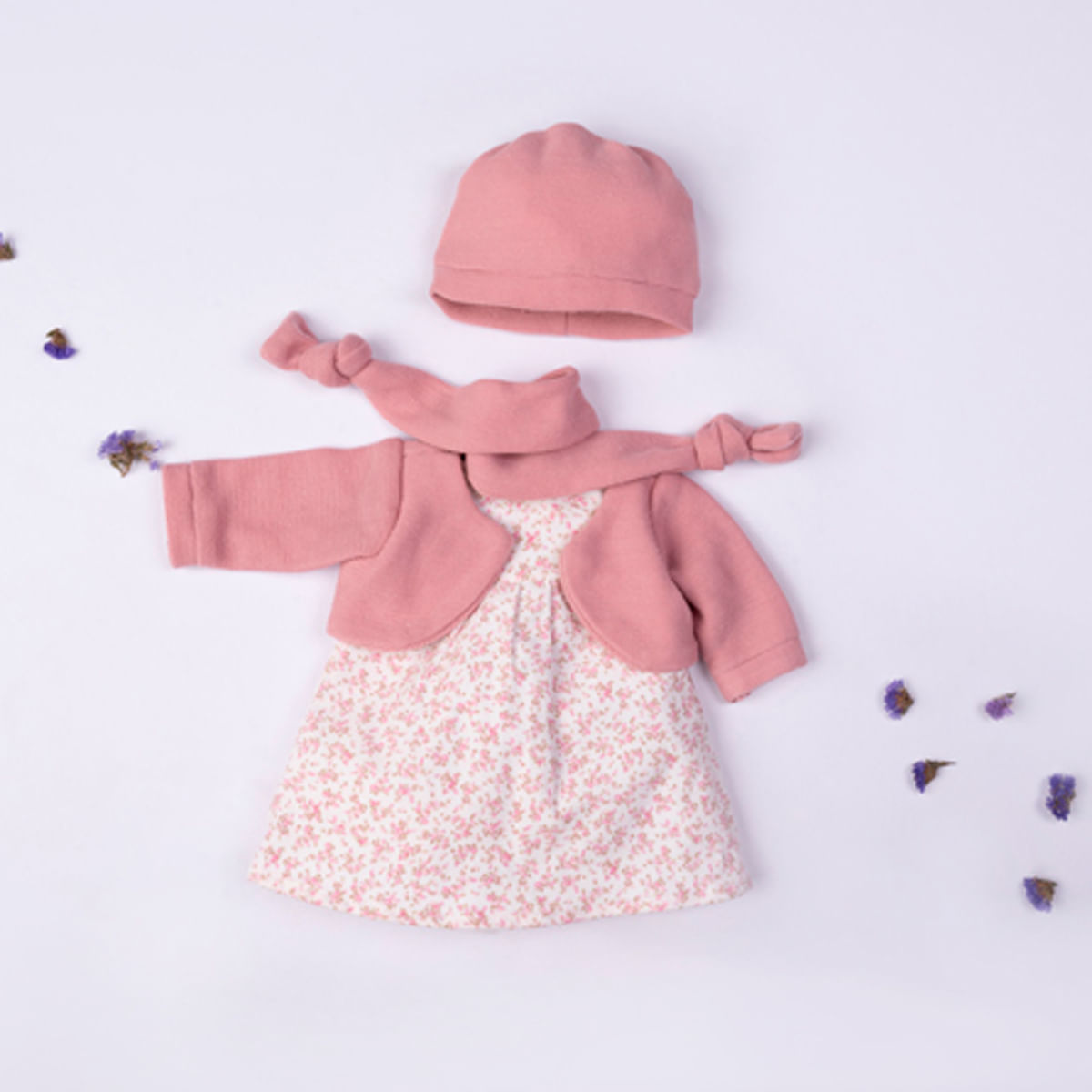 Un conjunto de ropa de bebé está sobre una superficie blanca. Incluye gorro, bufanda, chaqueta y vestido floral rosas. Flores secas dispersas.