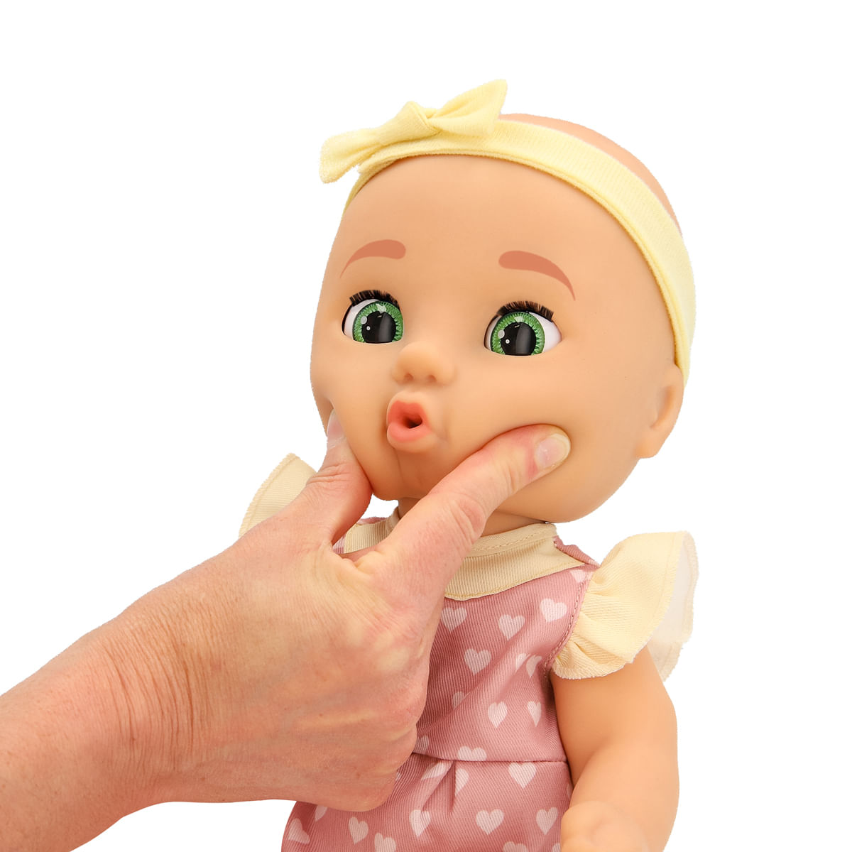 Una muñeca con ojos verdes y diadema amarilla tiene las mejillas pellizcadas por una mano. Lleva un vestido rosa con corazones blancos.