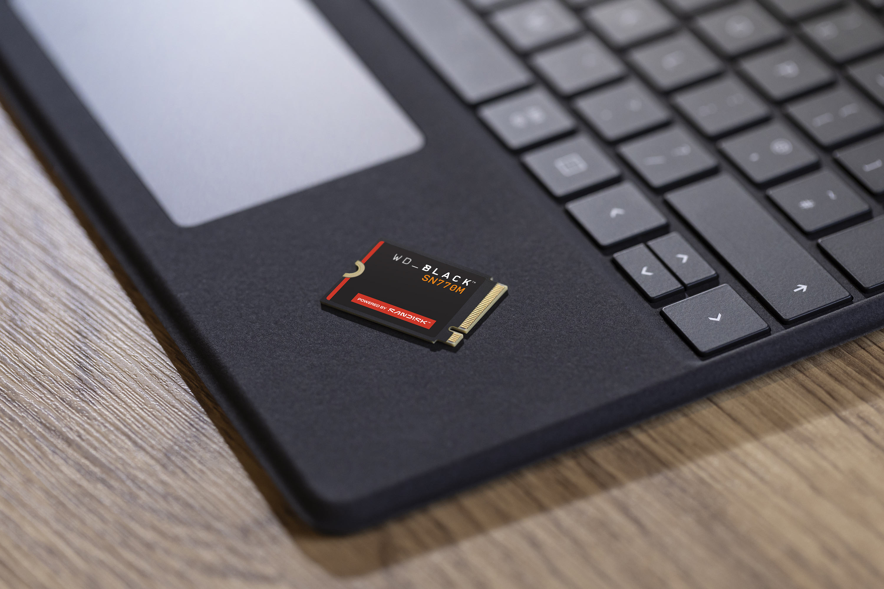 Dysk SSD leży na touchpadzie laptopa. Po prawej stronie klawiatura. Tło to drewno.