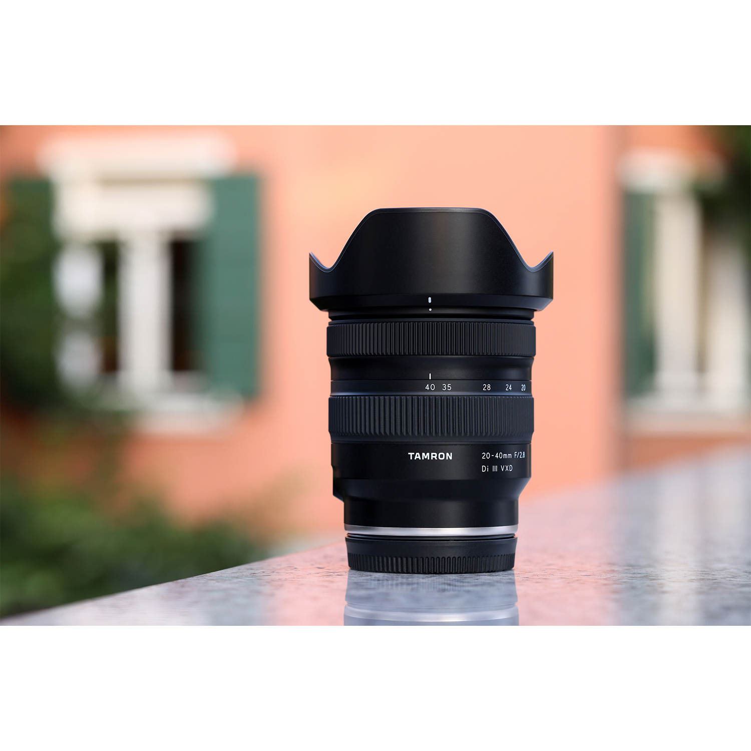 Ein schwarzes Tamron-Objektiv liegt auf einer reflektierenden Oberfläche. Dahinter ist ein verschwommenes Gebäude zu sehen.