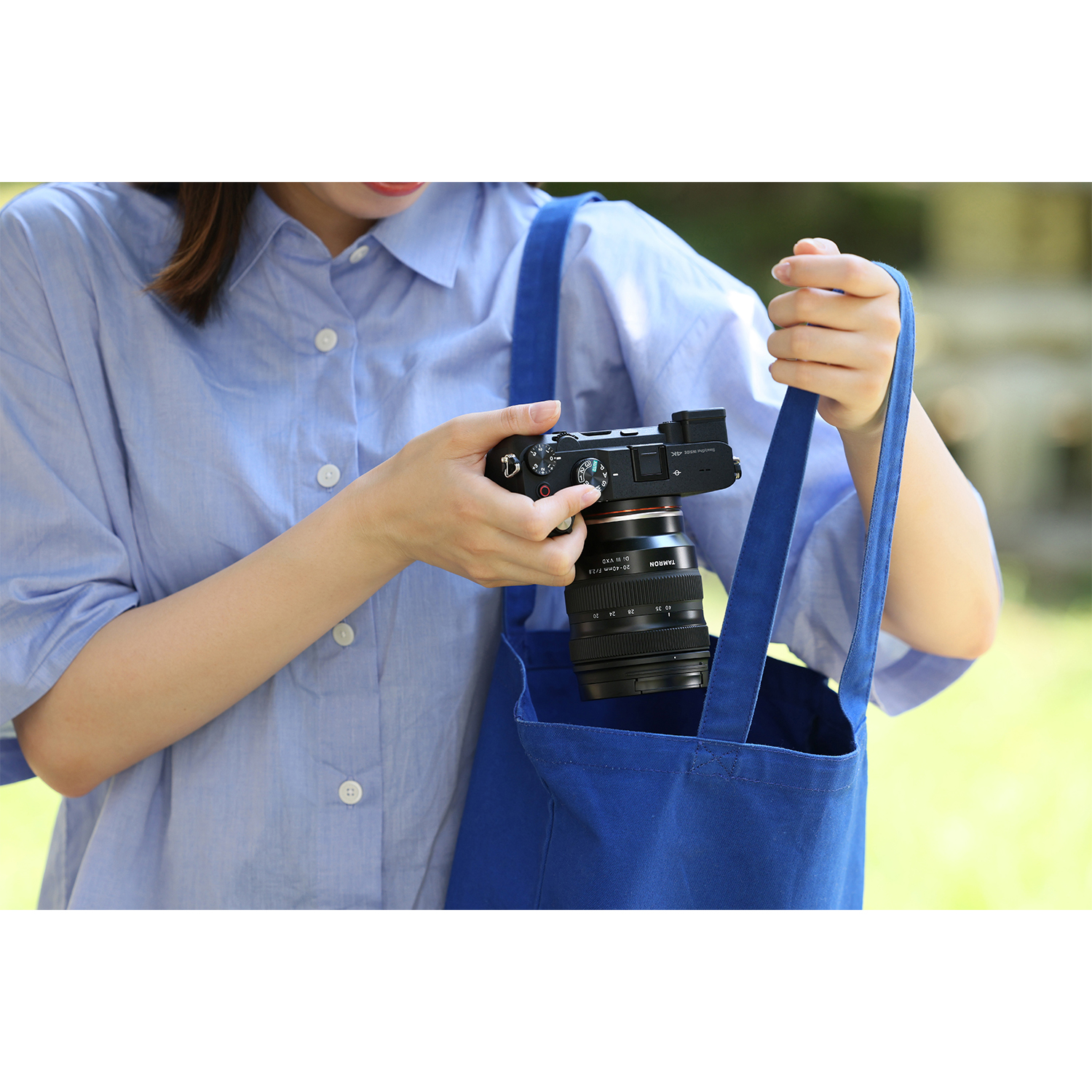Eine Person hält eine Kamera in einer blauen Tasche. Die Person trägt ein blaues Hemd. Grüner Hintergrund, Bokeh-Effekt.