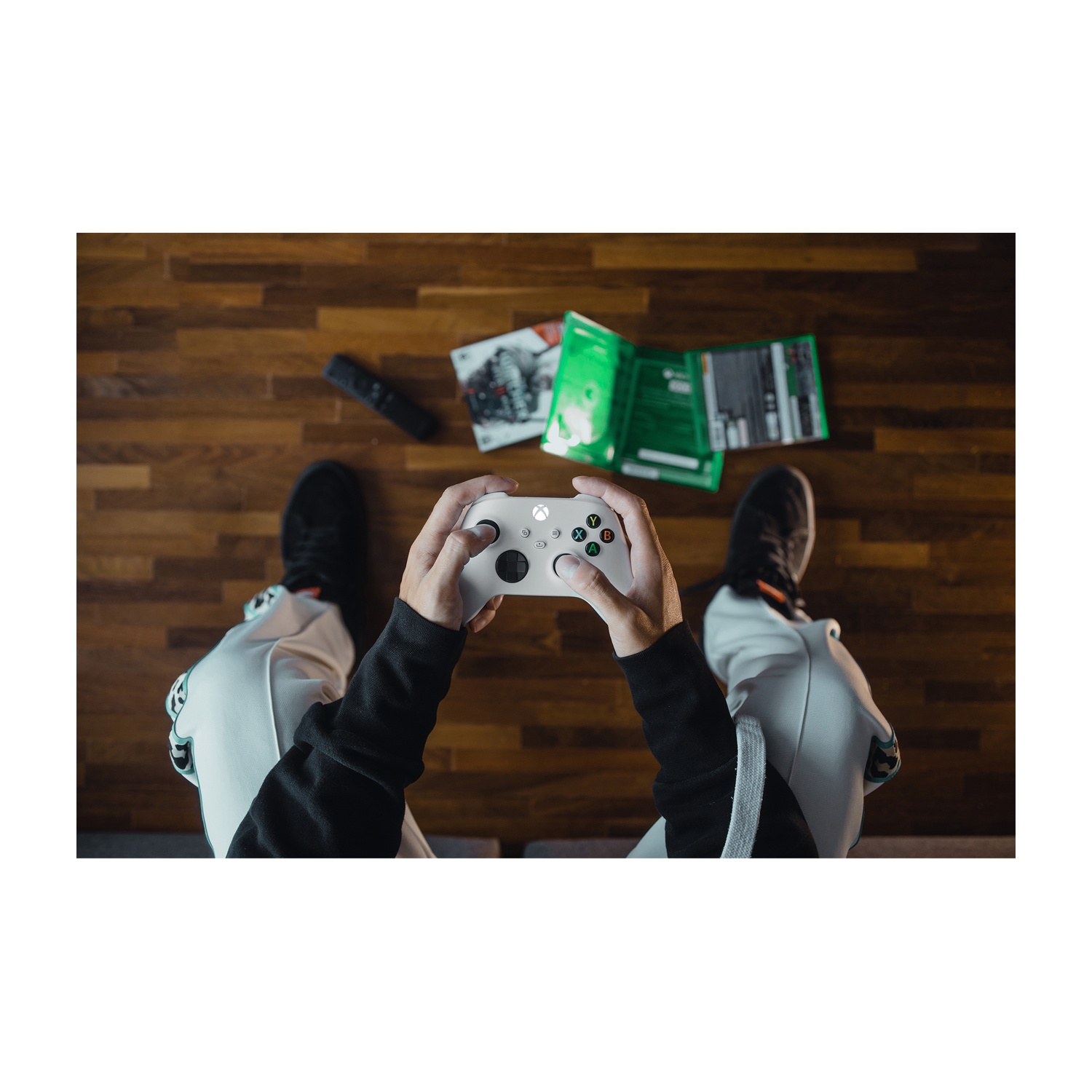 Een persoon houdt een witte Xbox-controller vast. Er liggen gamehandleidingen en een afstandsbediening op de houten vloer. De persoon draagt een witte broek.