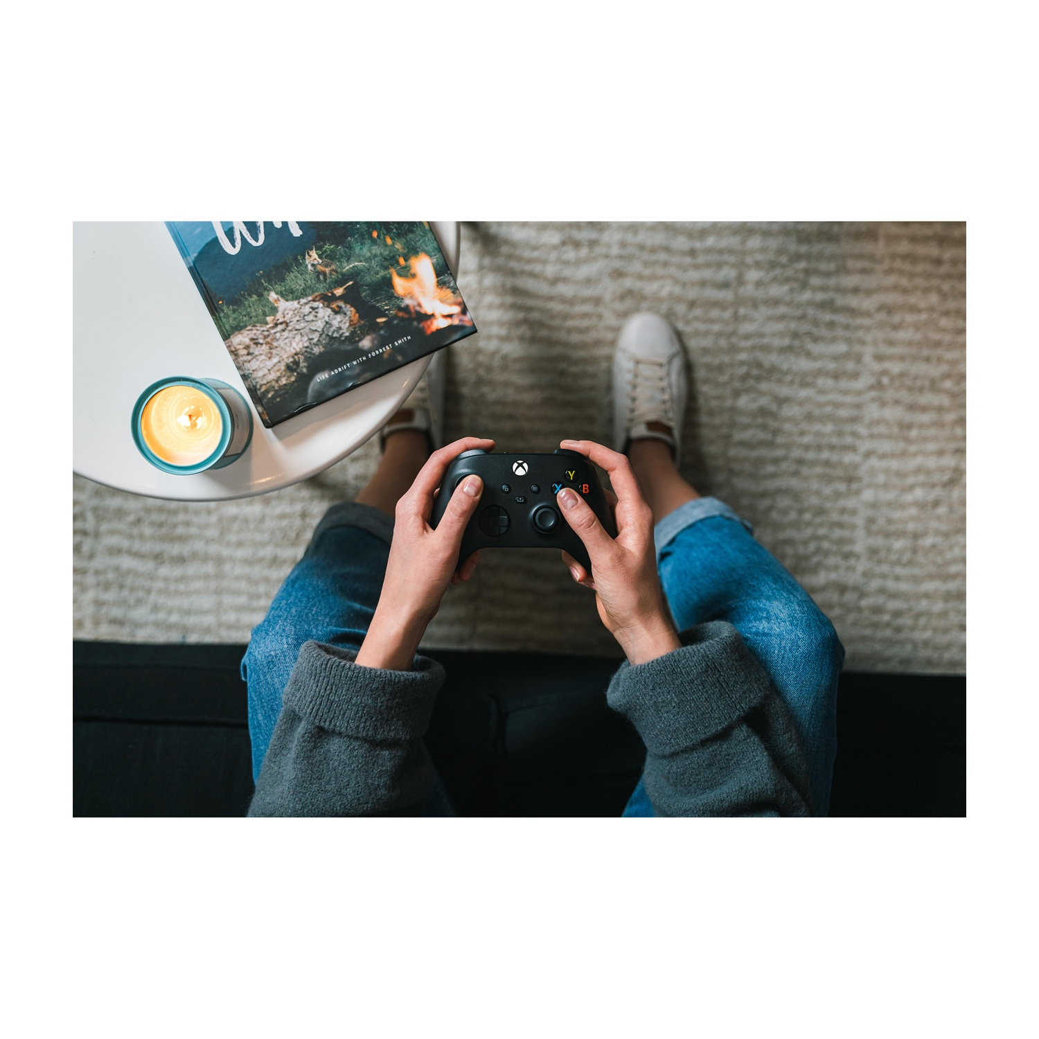 Osoba trzyma czarny kontroler Xbox. Na białym stole leży książka i świeca. Osoba ma na sobie dżinsy i białe buty.