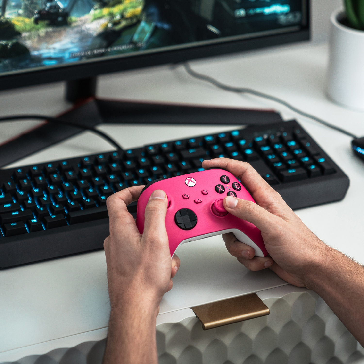 Een persoon houdt een roze Xbox-controller vast. Een zwart toetsenbord en monitor staan op de achtergrond, samen met een wit bureau.