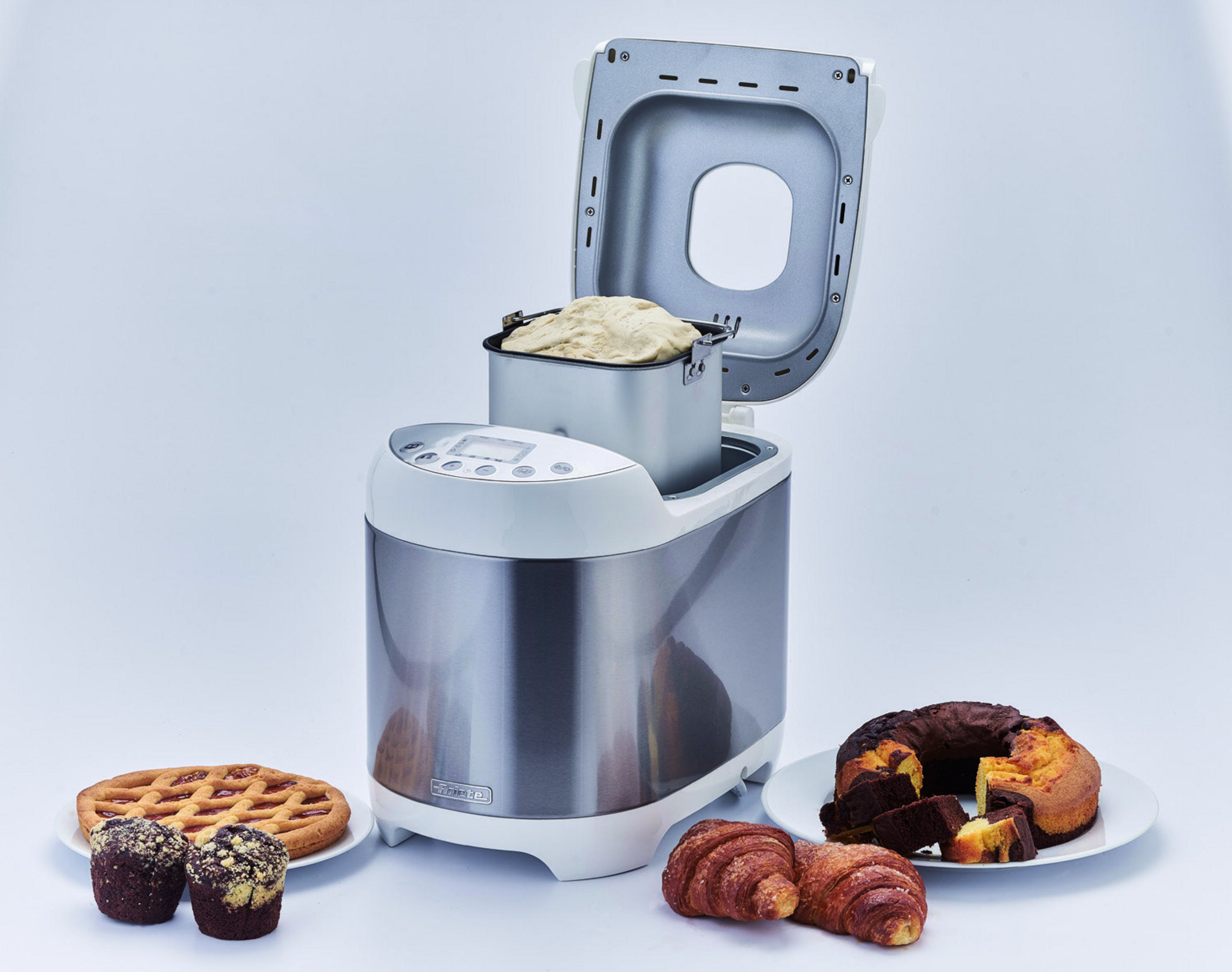 Brotmaschine mit offenem Deckel und Teig. Kuchen, Muffins und Croissants stehen auf dem Tisch.