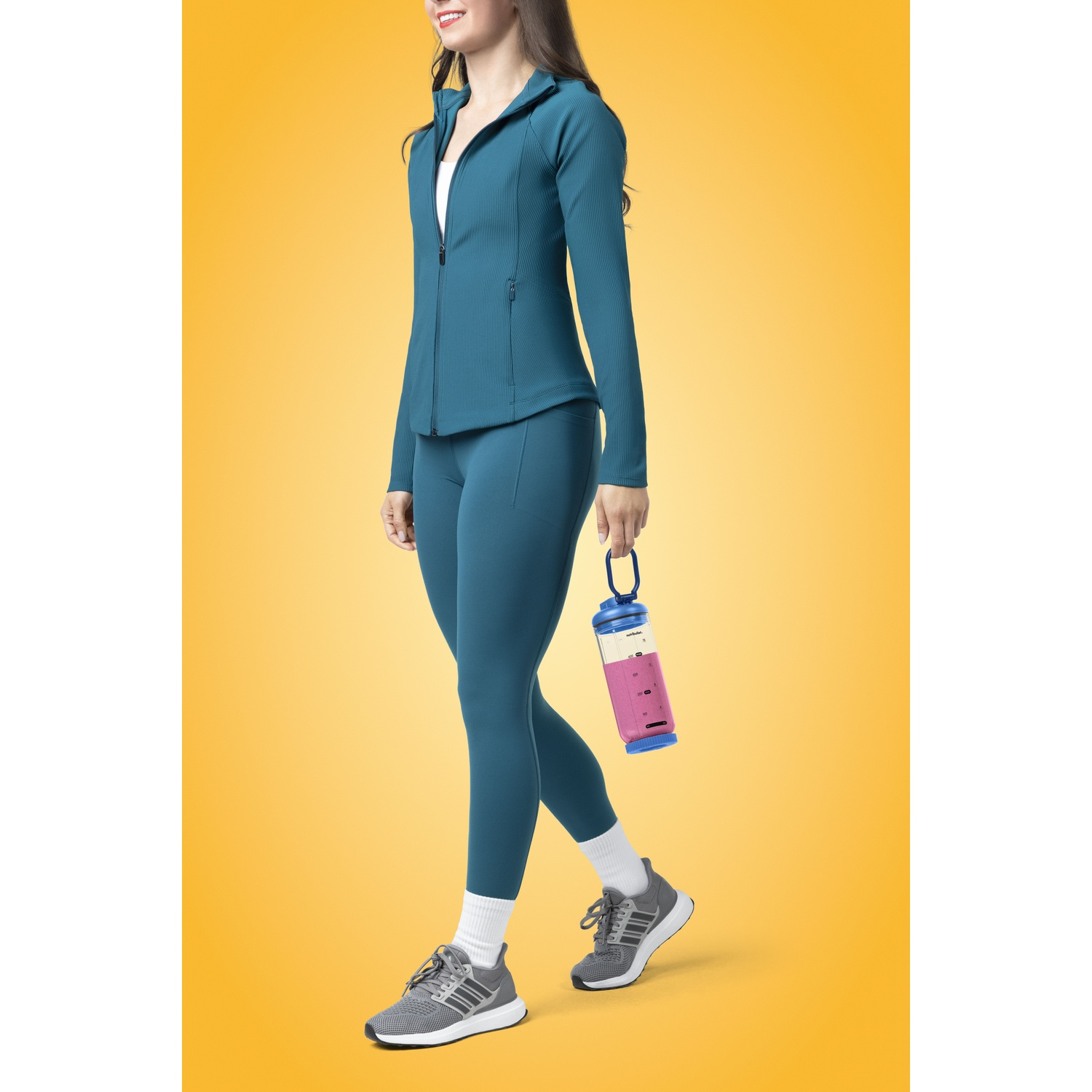 Frau in tealfarbener Trainingskleidung, hält einen rosa-blauen Shaker, geht auf gelbem Hintergrund mit grauen Schuhen.