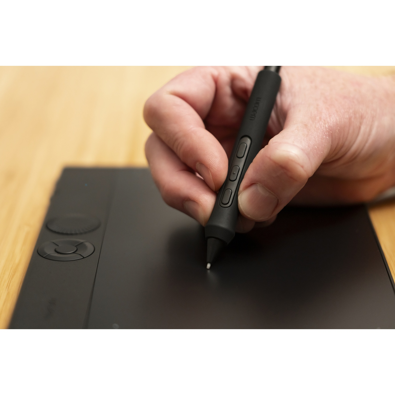 Eine Hand, die einen schwarzen Stift hält, benutzt ihn auf einem schwarzen Tablet. Der Hintergrund ist eine Holzoberfläche. Der Stift hat Knöpfe.
