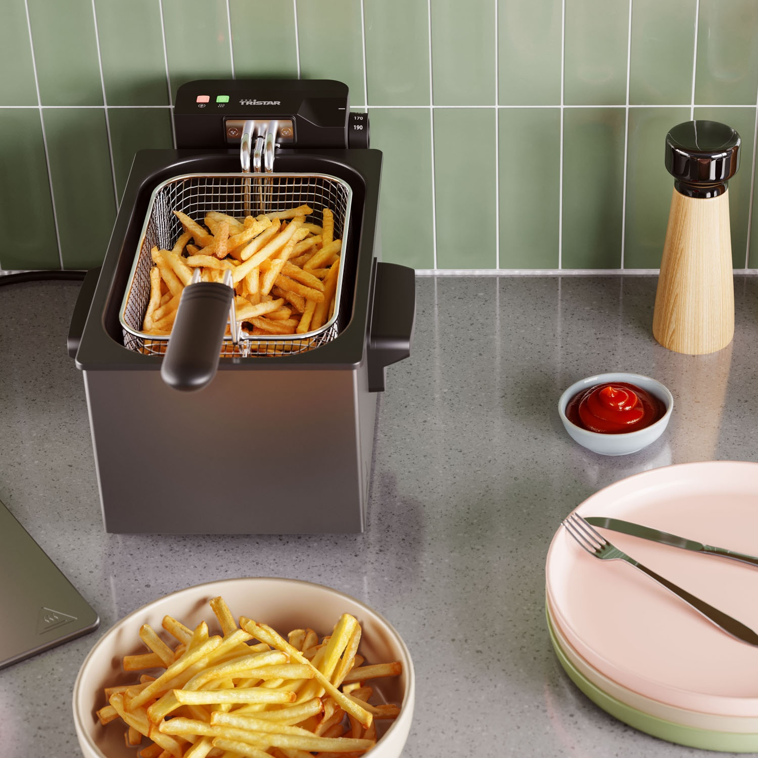Una friggitrice con patatine fritte, ketchup, piatti, saliera e una ciotola di patatine fritte su un piano di lavoro. Parete piastrellata verde.
