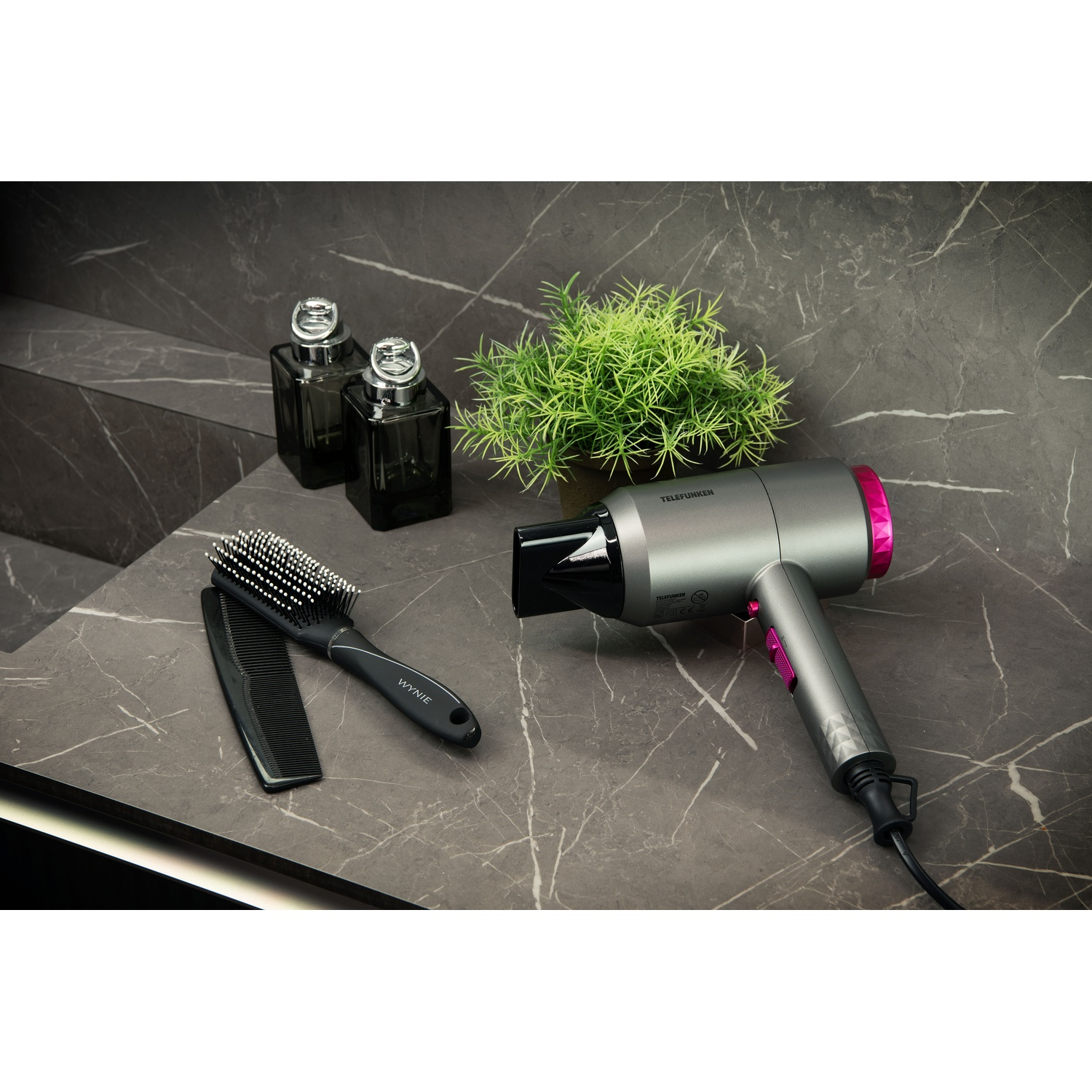 Szara suszarka do włosów z różowymi akcentami, szczotka do włosów, grzebień, mała zielona roślina i dwie ciemne butelki na marmurze.