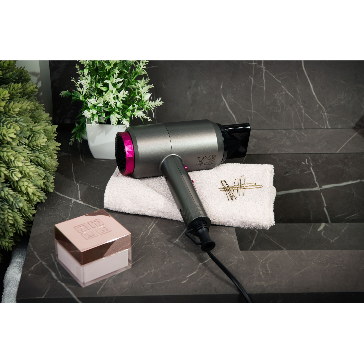 Szara suszarka do włosów z różowym akcentem leży na białym ręczniku. Szpilki do włosów obok. Kompakt do makijażu i rośliny w tle.