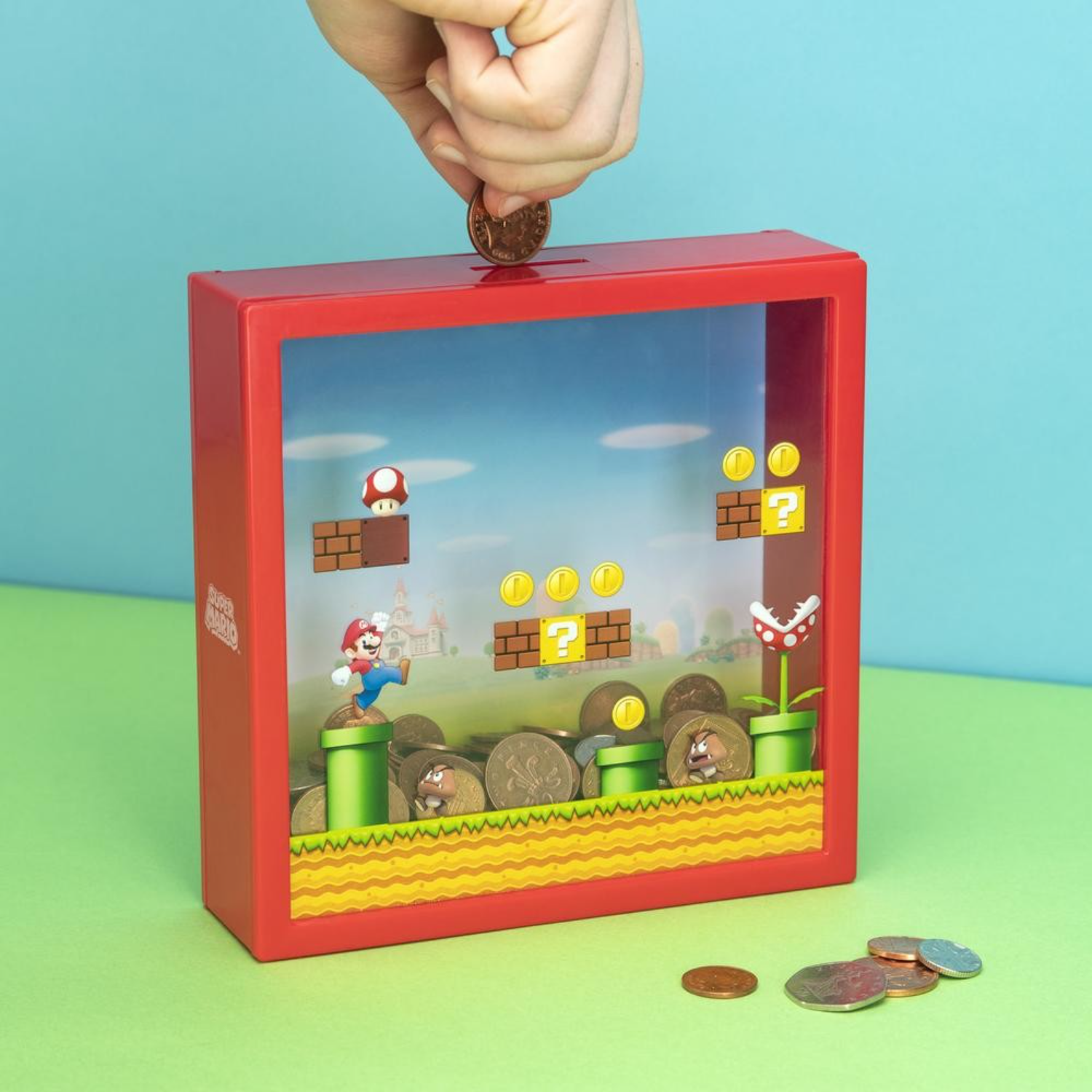 Eine Hand wirft eine Münze in ein rotes Super Mario-Sparschwein, das mit Münzen gefüllt ist und eine Mario-Szene zeigt.