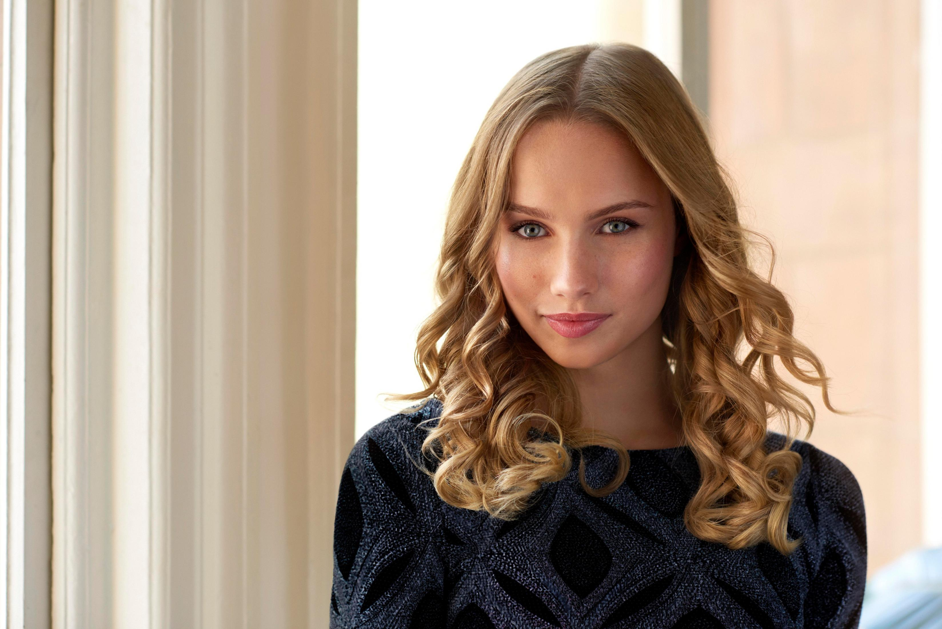 Kobieta z blond, kręconymi włosami i niebieskimi oczami patrzy na widza. Ma na sobie ciemny top. Tło to okno.