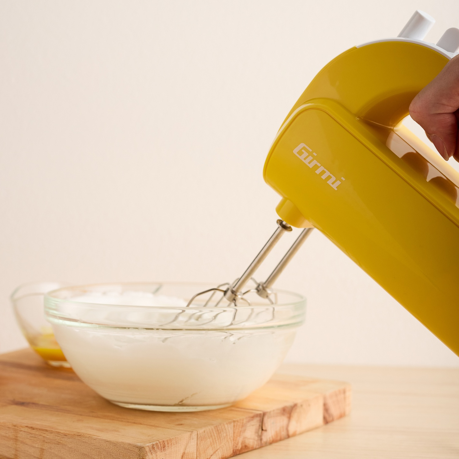 Un frullatore a mano giallo sta mescolando una sostanza bianca in una ciotola di vetro. Un'altra ciotola con liquido è sullo sfondo su una tavola di legno.