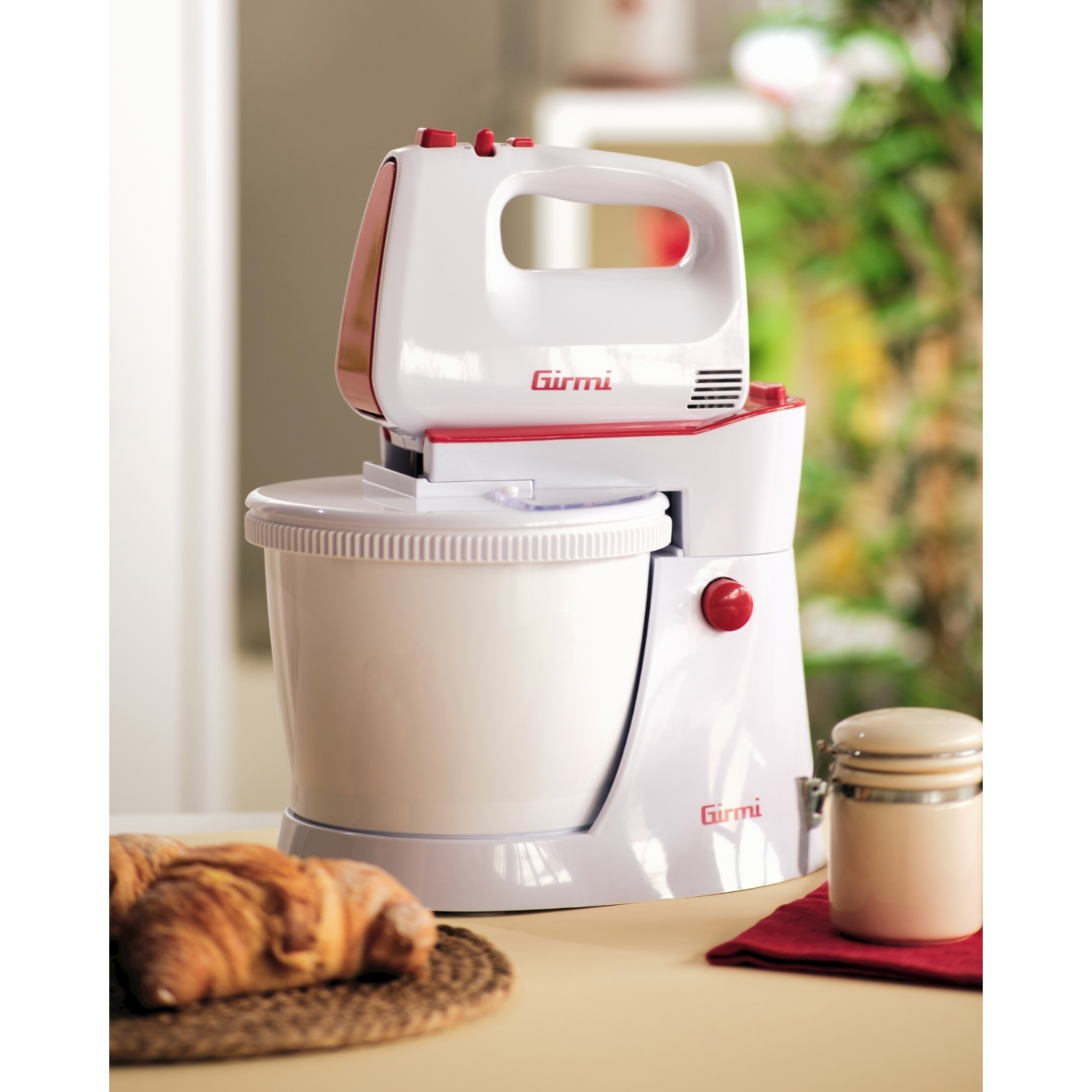 Un mixer Girmi bianco e rosso con ciotola, croissant e un barattolo su un tavolo. Sfondo sfocato di una cucina.
