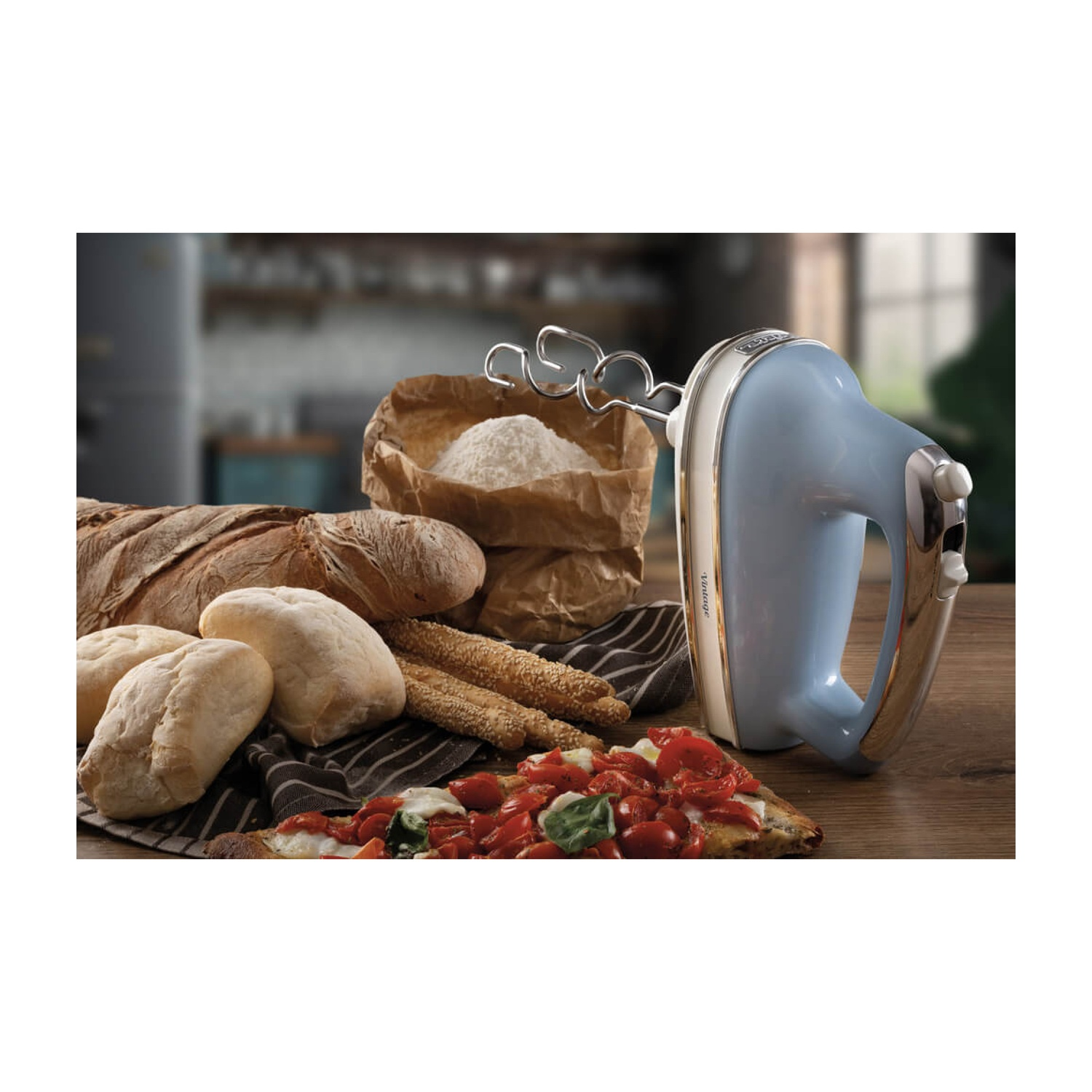 Ein blauer Handmixer mit Brot, Mehl und Pizza auf einem Holztisch. Der Hintergrund zeigt eine verschwommene Küchenszene.