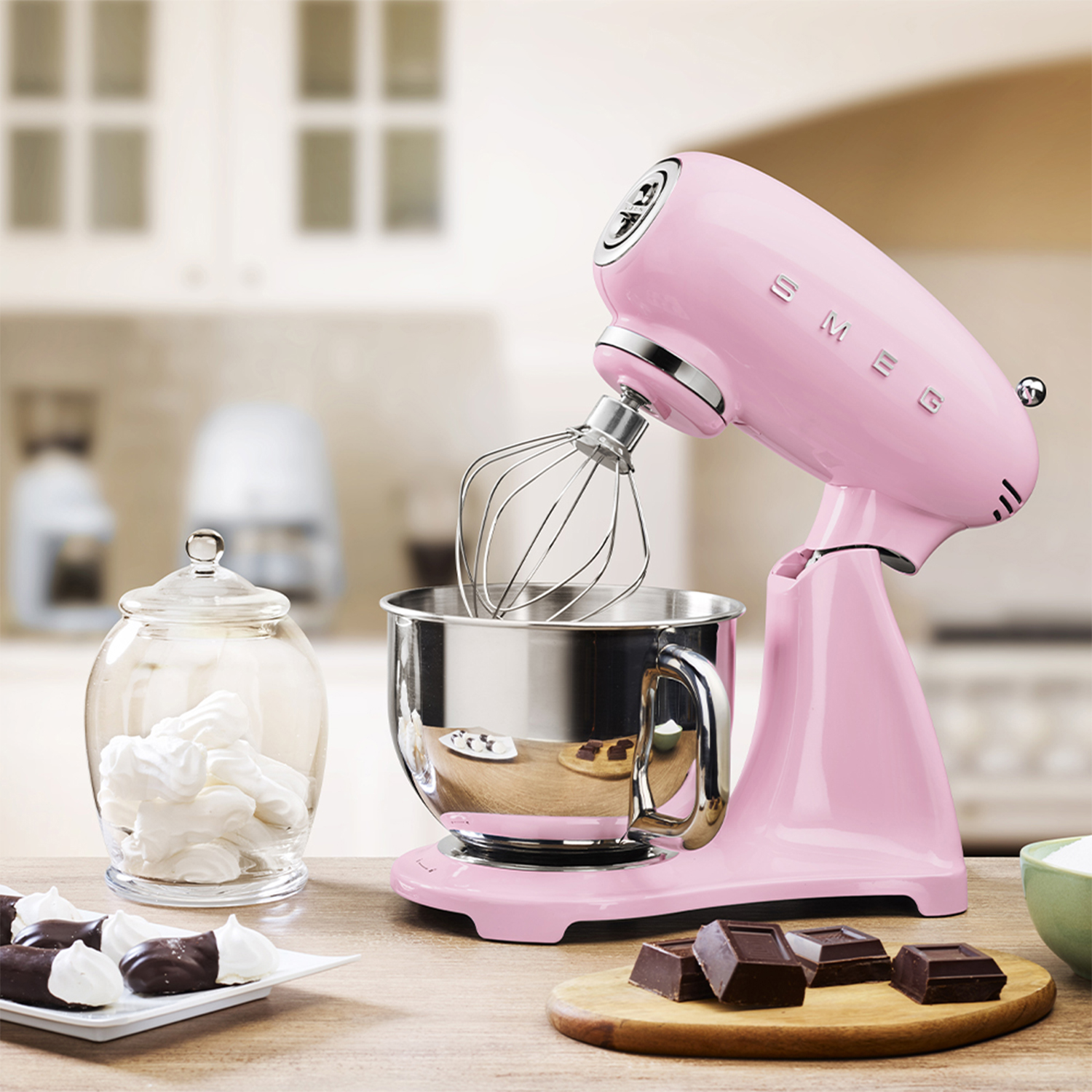 Un'impastatrice Smeg rosa con frusta, un barattolo di meringhe e pezzi di cioccolato su una superficie di legno.