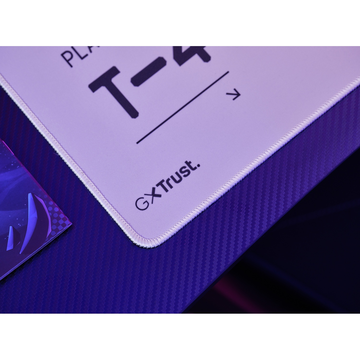 Gros plan d'un tapis de souris Trust GX. Il comporte le texte T- et une flèche. La surface est blanche et sur une surface bleu foncé.