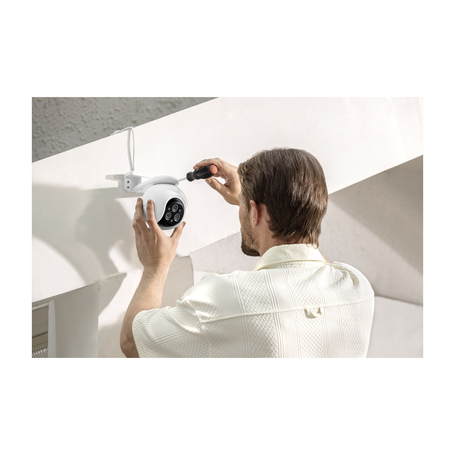 Ein Mann installiert eine weiße Überwachungskamera an einer weißen Wand. Er benutzt einen Schraubendreher. Die Kamera hat schwarze Linsen.