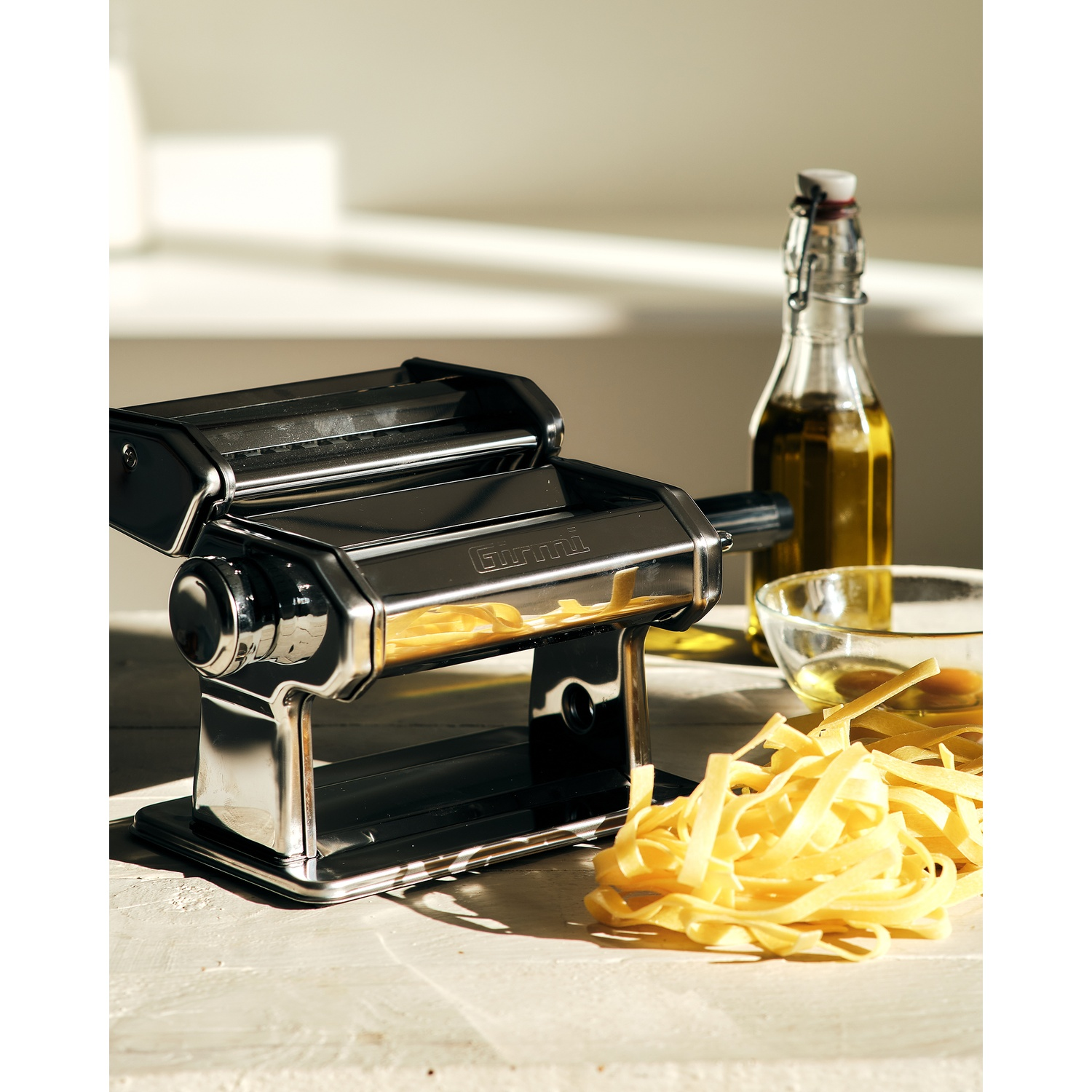 Una macchina per la pasta con pasta fresca. Bottiglia di olio d'oliva e ciotola visibili. Luce solare su legno bianco.