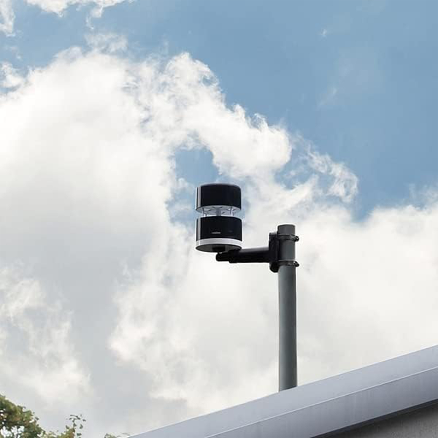 A weather station on a pole against a cloudy sky. The station is black and white, mounted on a gray pole. The roof is visible.