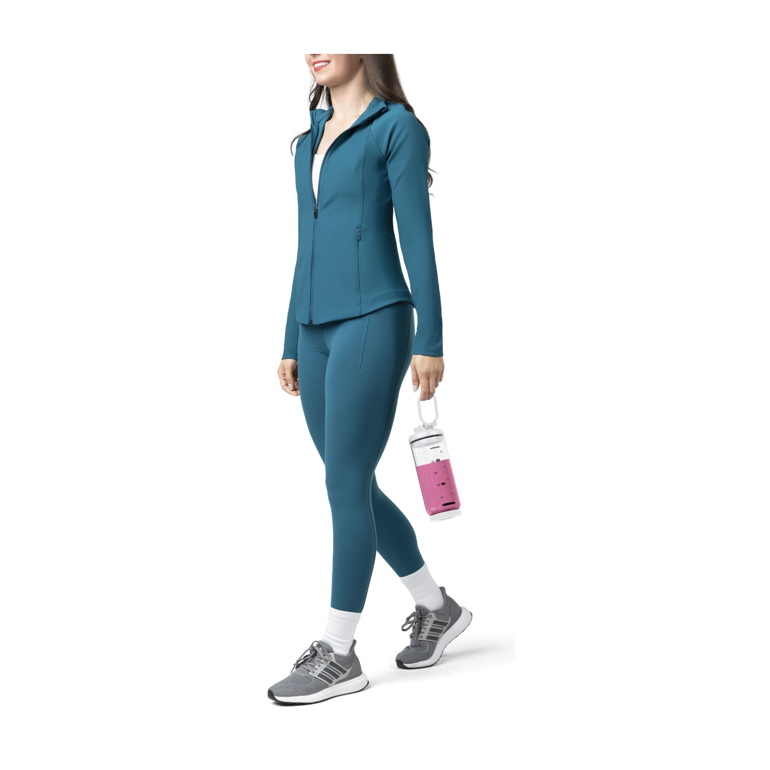 Eine Frau in tealfarbener Sportkleidung geht und hält eine rosa-weiße Flasche. Sie trägt graue Turnschuhe und weiße Socken vor weißem Hintergrund.