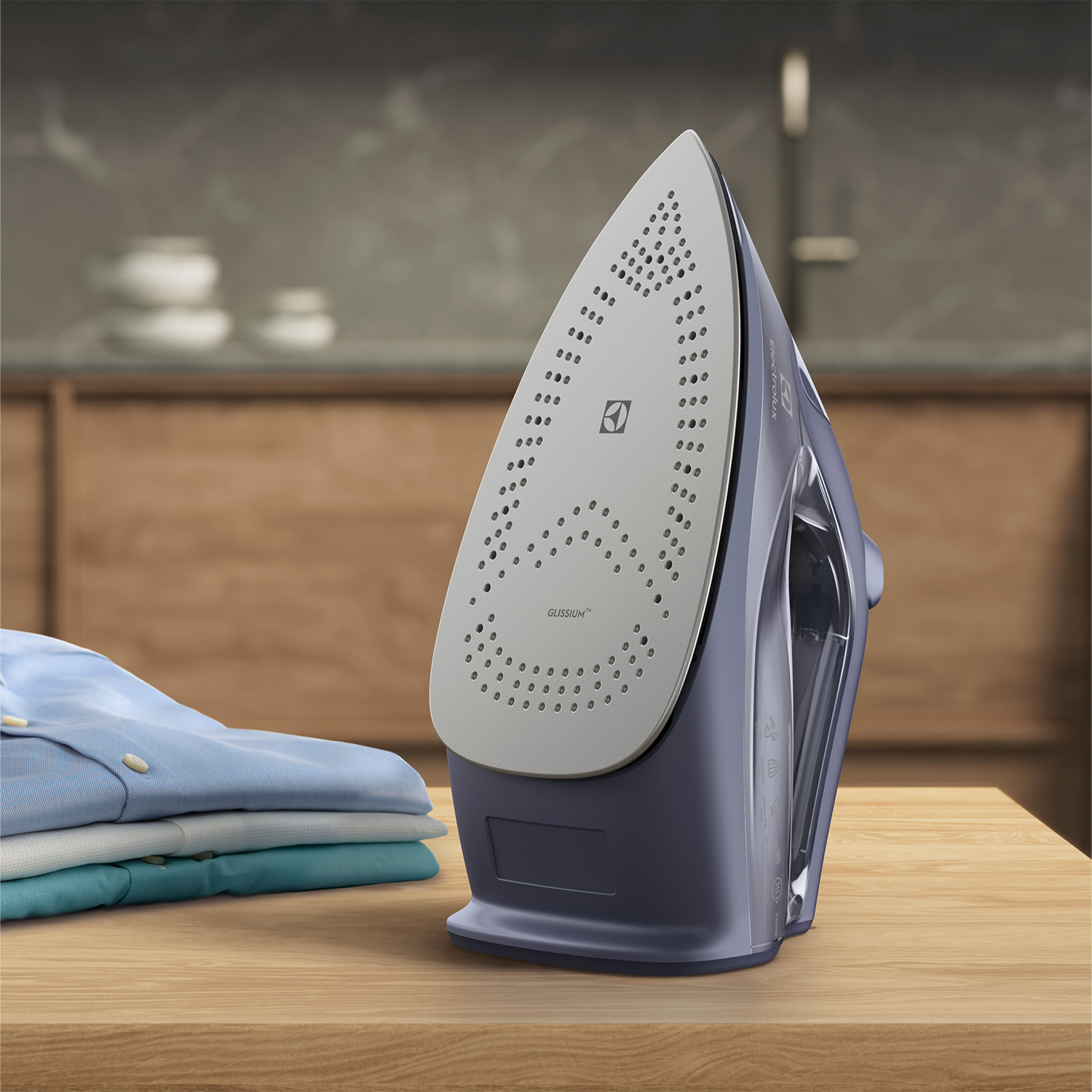 Niebieskie żelazko na drewnianym stole obok stosu złożonych koszul. W tle szafki i rozmyta bateria.