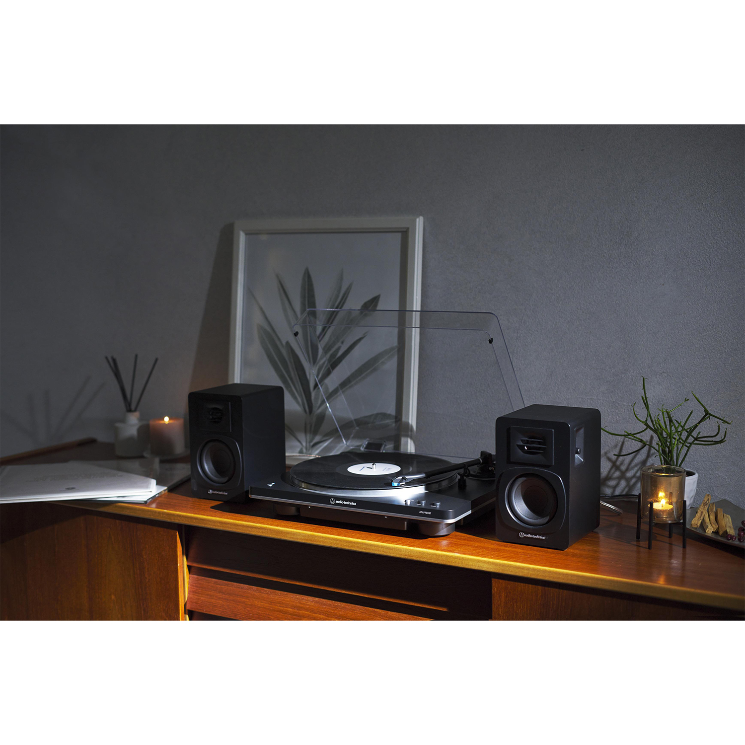 Gramofon z dwoma głośnikami na drewnianej szafce, obraz w ramie na ścianie, świeca i roślina po prawej stronie.
