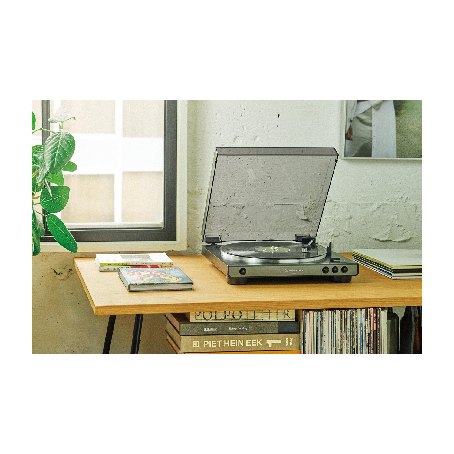 Gramofon z przezroczystą pokrywą stoi na drewnianym stole z książkami. W tle okno i rośliny. Płyty winylowe są widoczne.