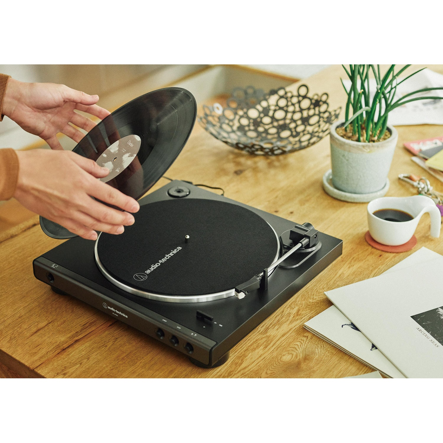 Osoba umieszcza płytę winylową na czarnym gramofonie. Filiżanka kawy, roślina i płyty znajdują się na drewnianym stole.