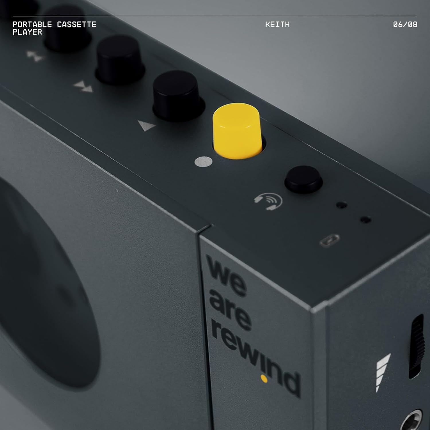 Un lecteur de cassettes portable gris foncé avec des boutons noirs et une commande jaune. Le texte 'we are rewind' est visible.