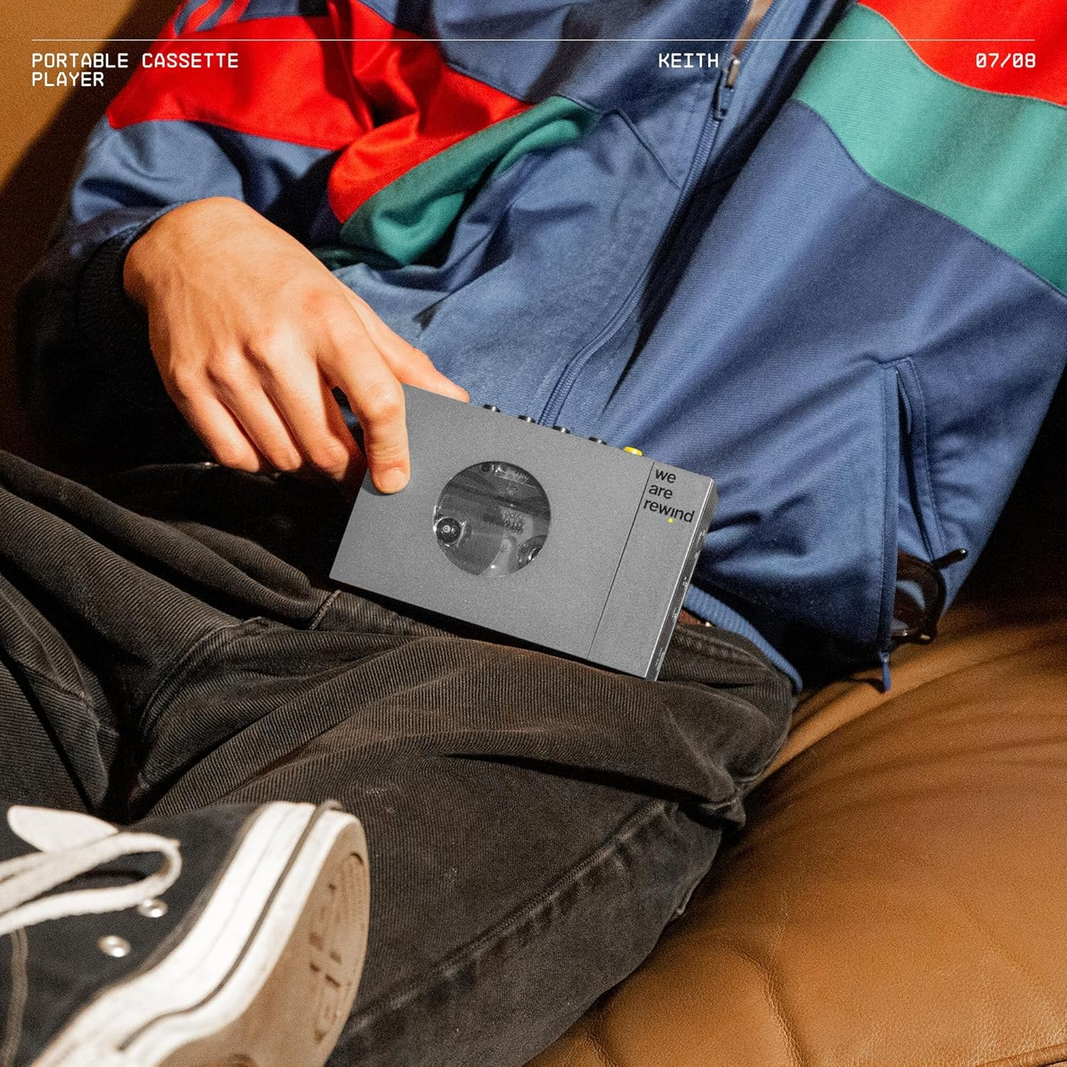 Personne tenant un lecteur de cassettes gris. Porte une veste bleue et un pantalon noir. Assis sur un canapé marron. Baskets blanches au premier plan.