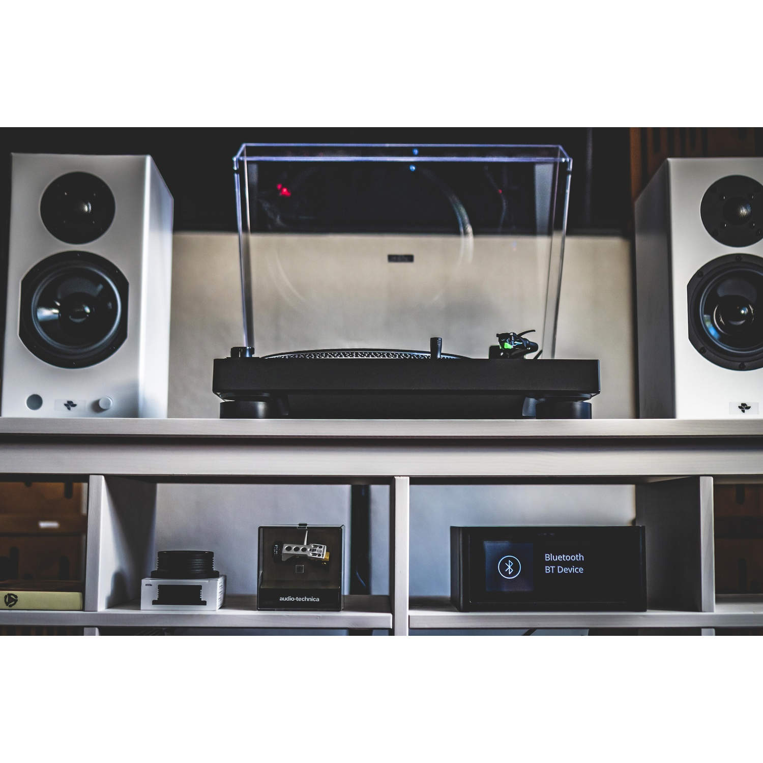 Gramofon stoi na półce między dwoma głośnikami. Poniżej sprzęt audio jest ułożony. Tło to ściana.