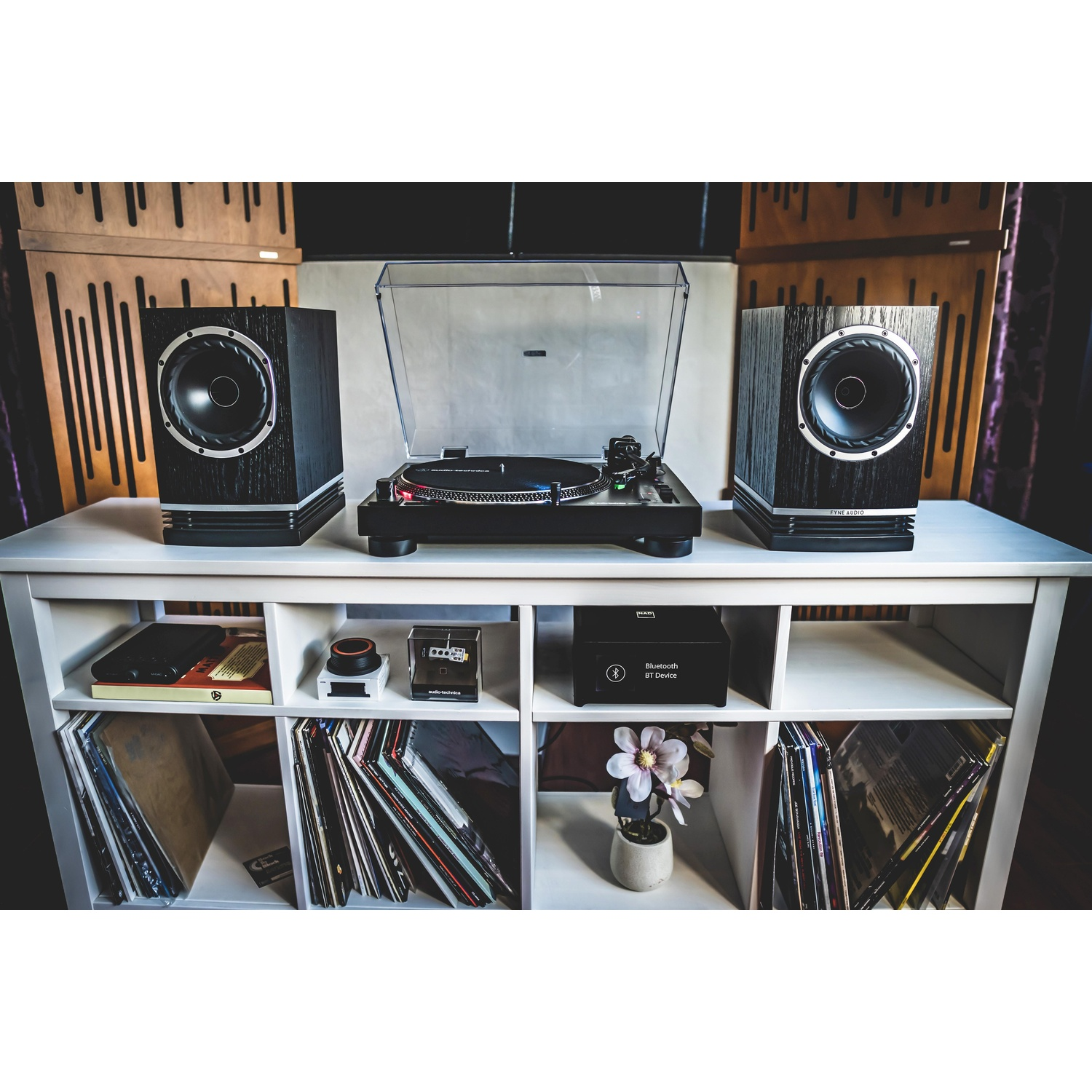 Gramofon z dwoma głośnikami na białej półce. Na półce znajdują się płyty. Obecne są również urządzenie Bluetooth i kwiat.