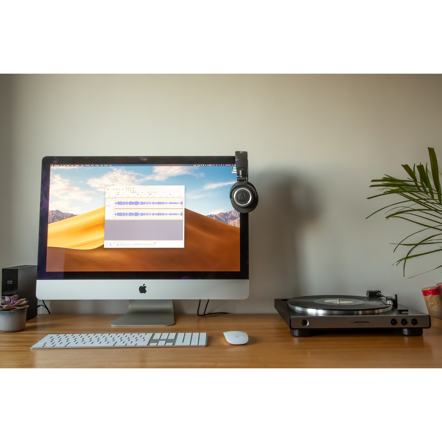 Biurko z komputerem, słuchawkami i gramofonem. Na drewnianej powierzchni znajduje się również sukulent i mysz.