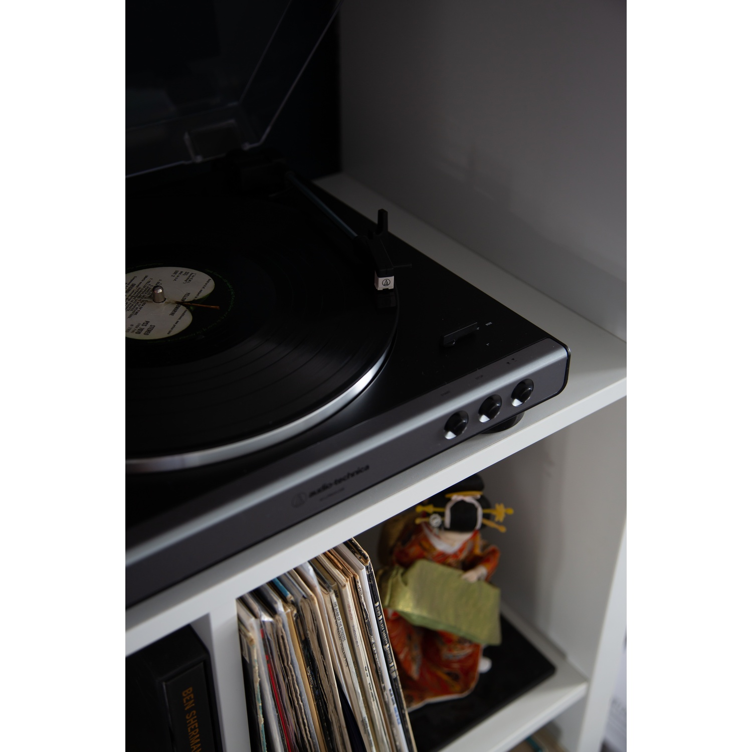 Gramofon z czarną płytą winylową stoi na białej półce. Widoczna jest również japońska lalka i albumy płytowe.