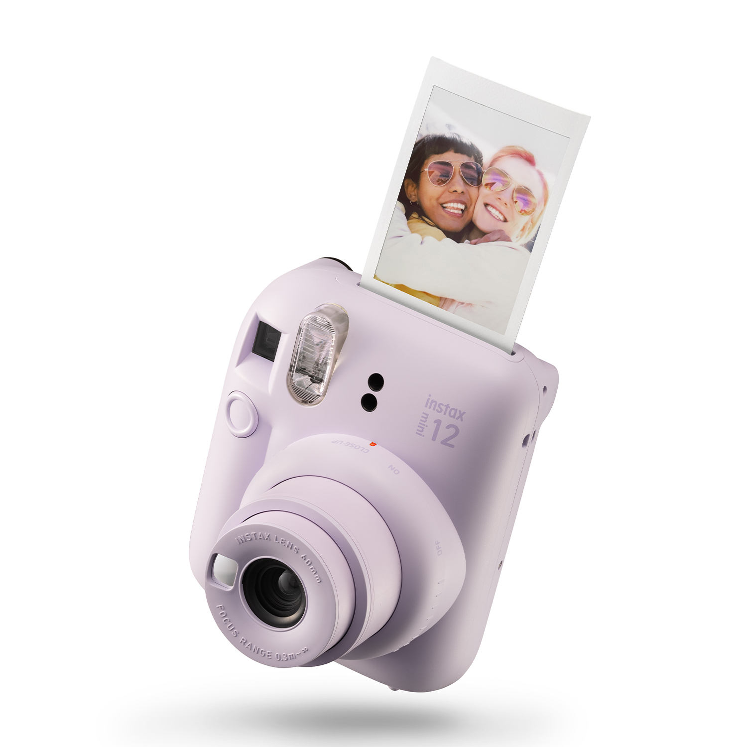Eine lavendelfarbene Instax Mini 12-Kamera ist abgebildet, aus der ein Foto von zwei Frauen herauskommt. Die Kamera hat ein Objektiv und Blitzlicht.