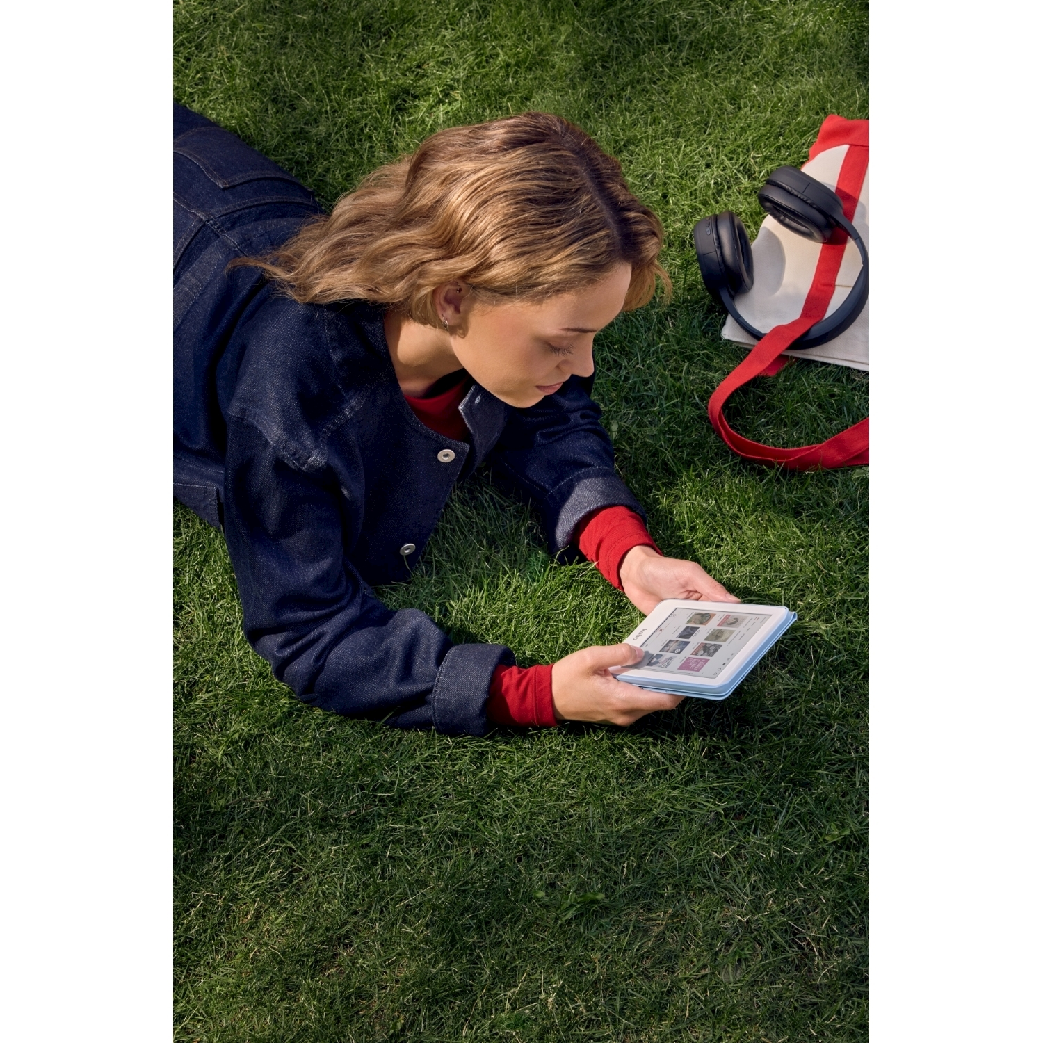 Kobieta leży na trawie, czytając tablet. Ma na sobie dżinsową kurtkę i czerwoną koszulę. Obok niej są słuchawki i torba.