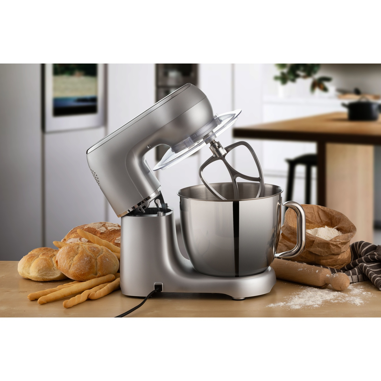 Ein silberner Küchenmixer mit Schüssel steht auf einer Holzoberfläche. Brot und Mehl sind in der Nähe. Im Hintergrund ist eine Küche zu sehen.