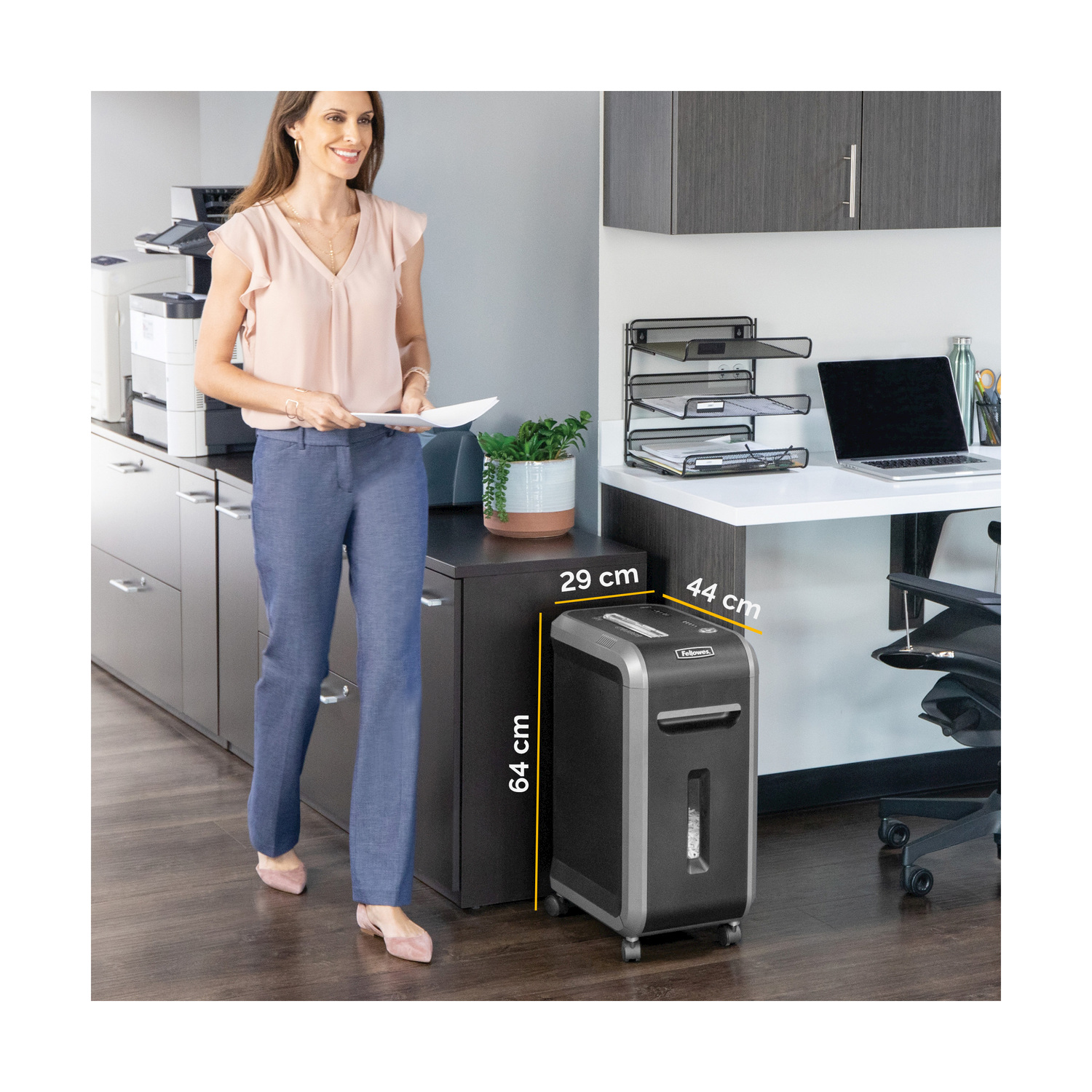 Kobieta w biurze trzyma papier, stoi przy niszczarce. Biurko z laptopem, tackami. Ciemne szafki, widoczna drukarka. Drewniana podłoga.