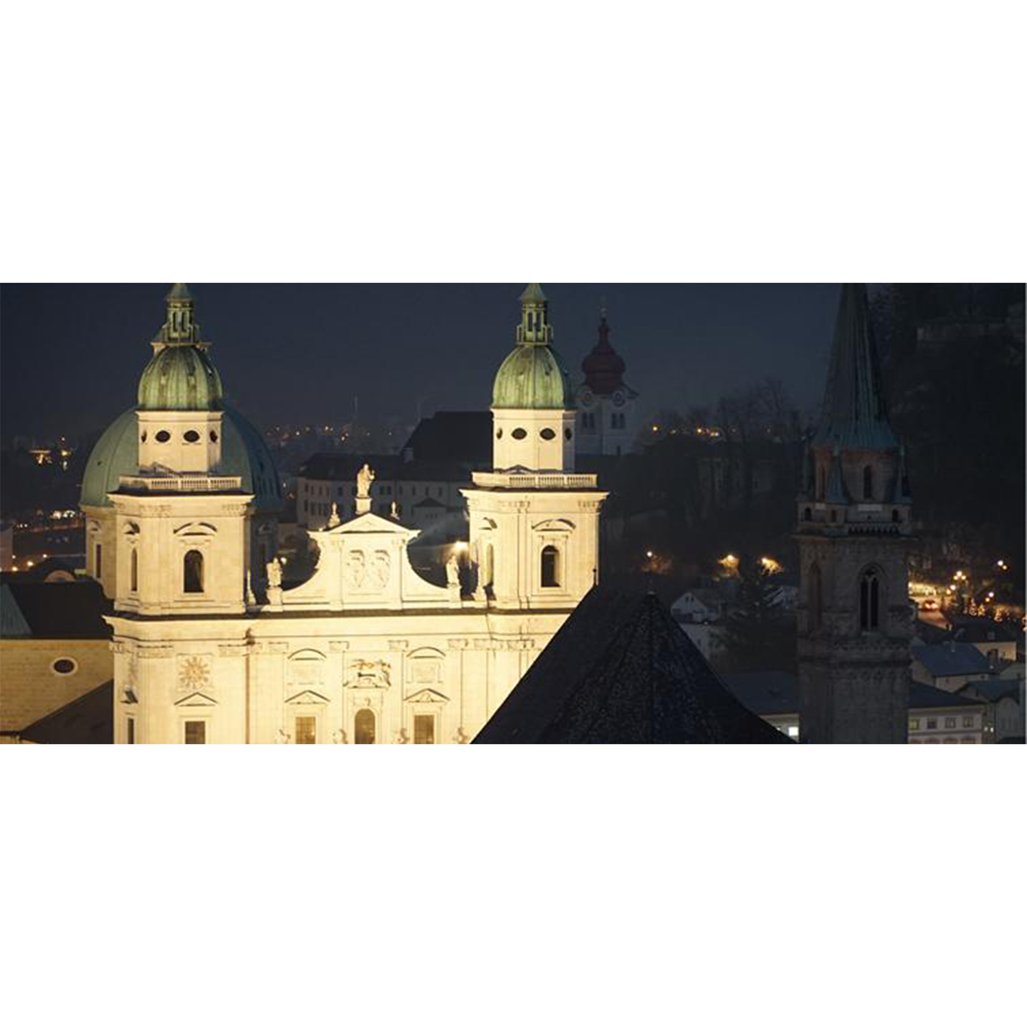 Vista nocturna de una ciudad con edificios iluminados. Destacan cúpulas y agujas de iglesias con luces a lo lejos. El cielo es oscuro.