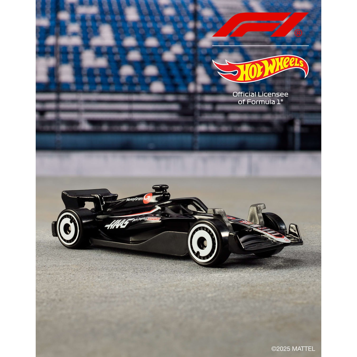 Czarny samochód Haas F1 Hot Wheels stoi na szarym betonie. Tło to rozmyty stadion z niebieskimi siedzeniami. Widoczne są loga.
