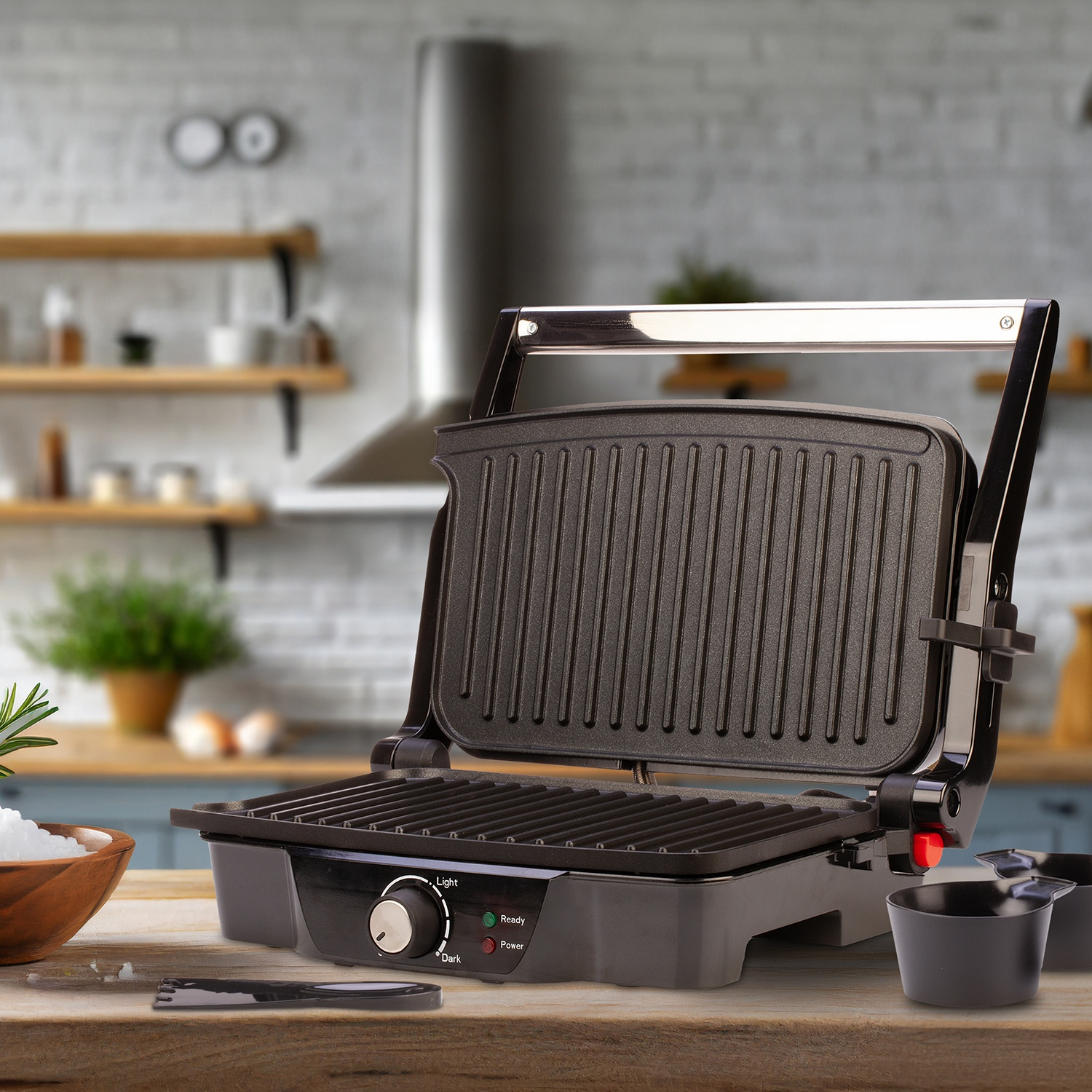 Una parrilla negra está abierta sobre una mesa de madera. Un cuenco de sal, un tenedor y tazas medidoras están delante. Cocina en el fondo.