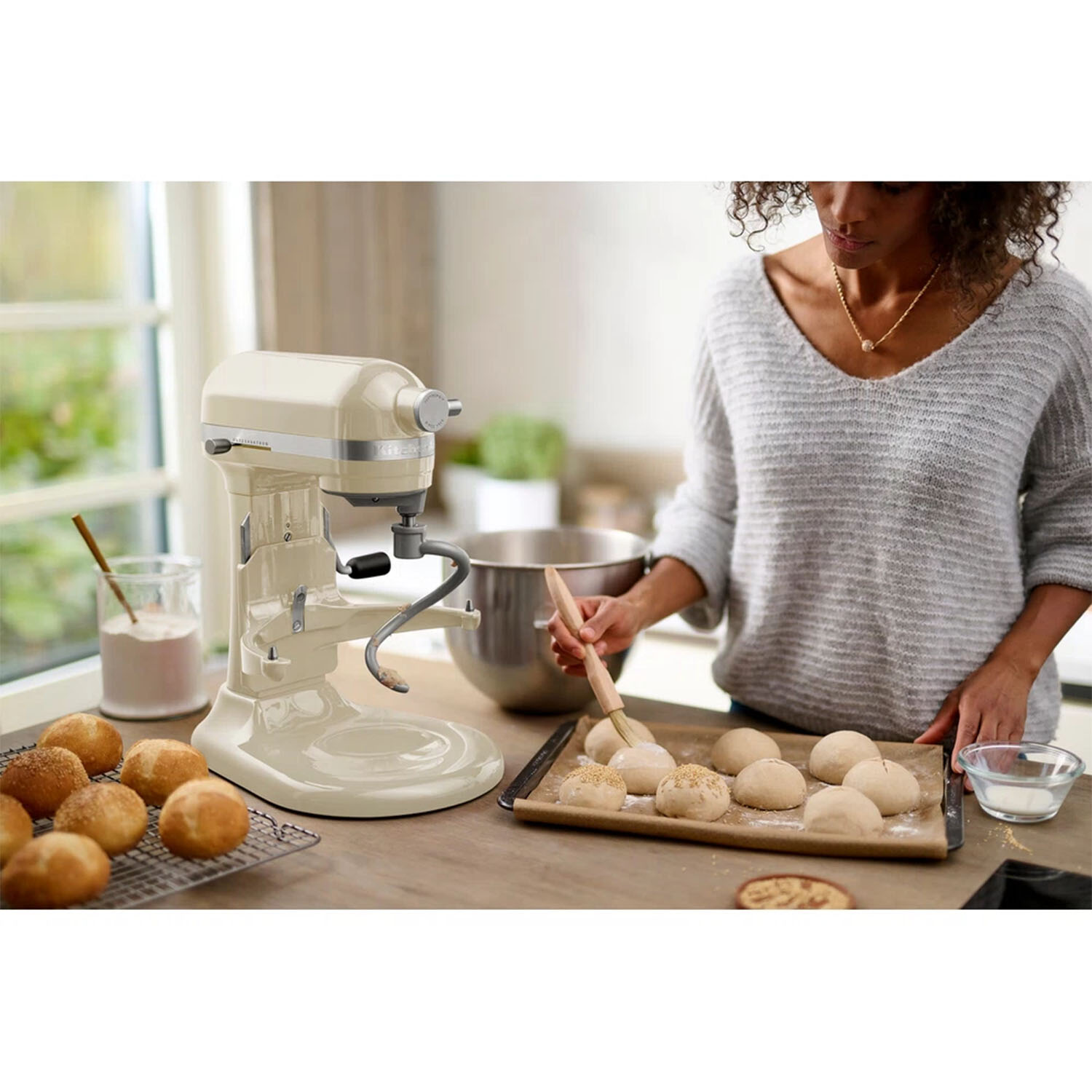 Eine Frau bestreicht Teig auf einem Backblech. Eine Küchenmaschine, Brötchen und Zutaten stehen auf der Theke.