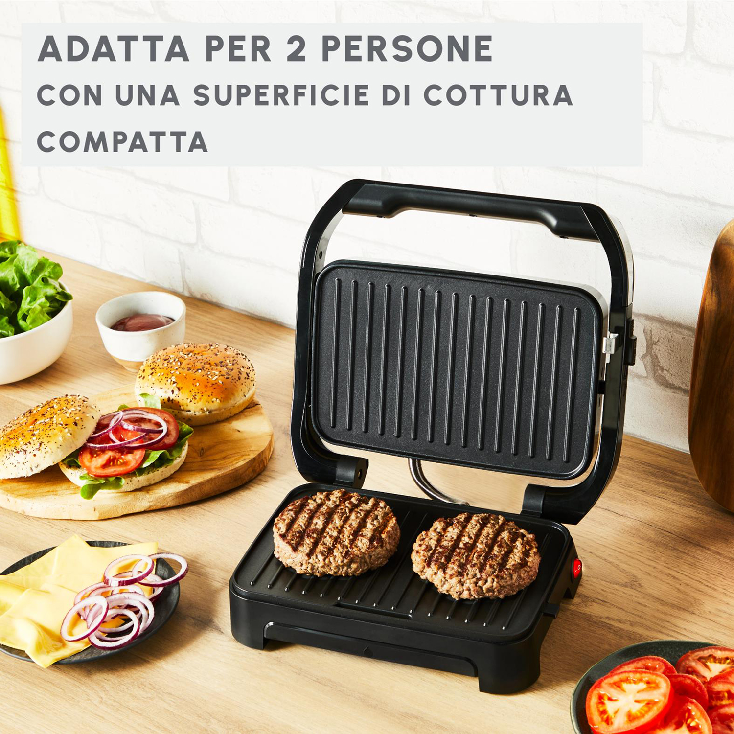 Offener Grill mit zwei bratenen Burgern. Burgerbrötchen, Käse, Tomaten und Zwiebeln auf dem Tisch. Text: Adatta per 2 persone.