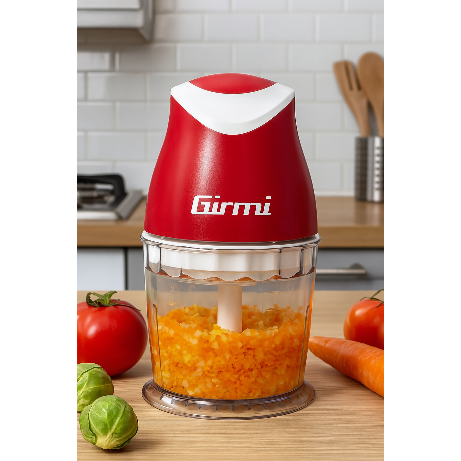 Un picador de alimentos Girmi rojo y blanco con verduras picadas dentro. Tomates, zanahorias y coles de Bruselas en la mesa.