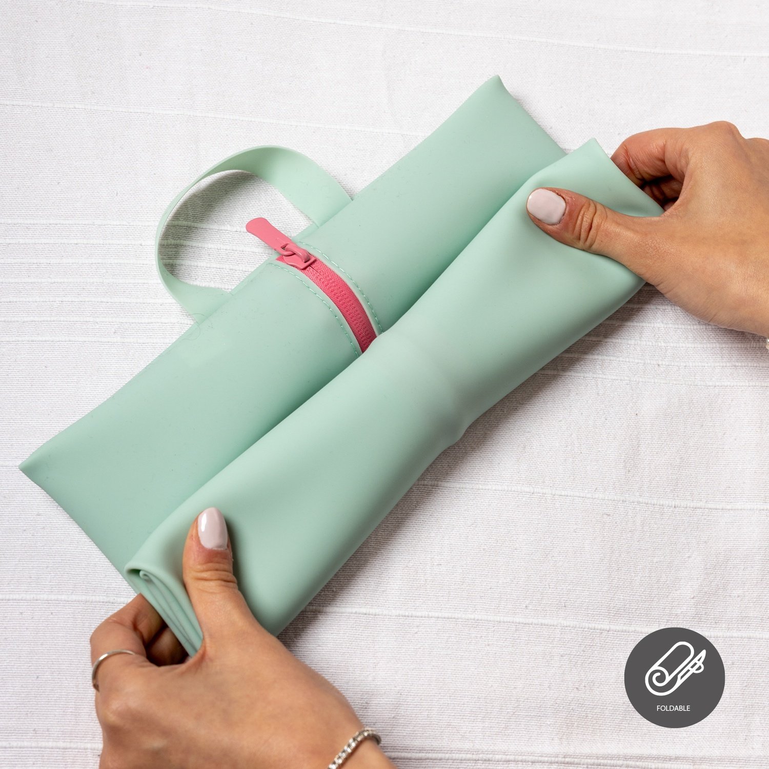 Manos desplegando una bolsa verde menta plegable con cremallera rosa. Está sobre una superficie blanca texturizada.