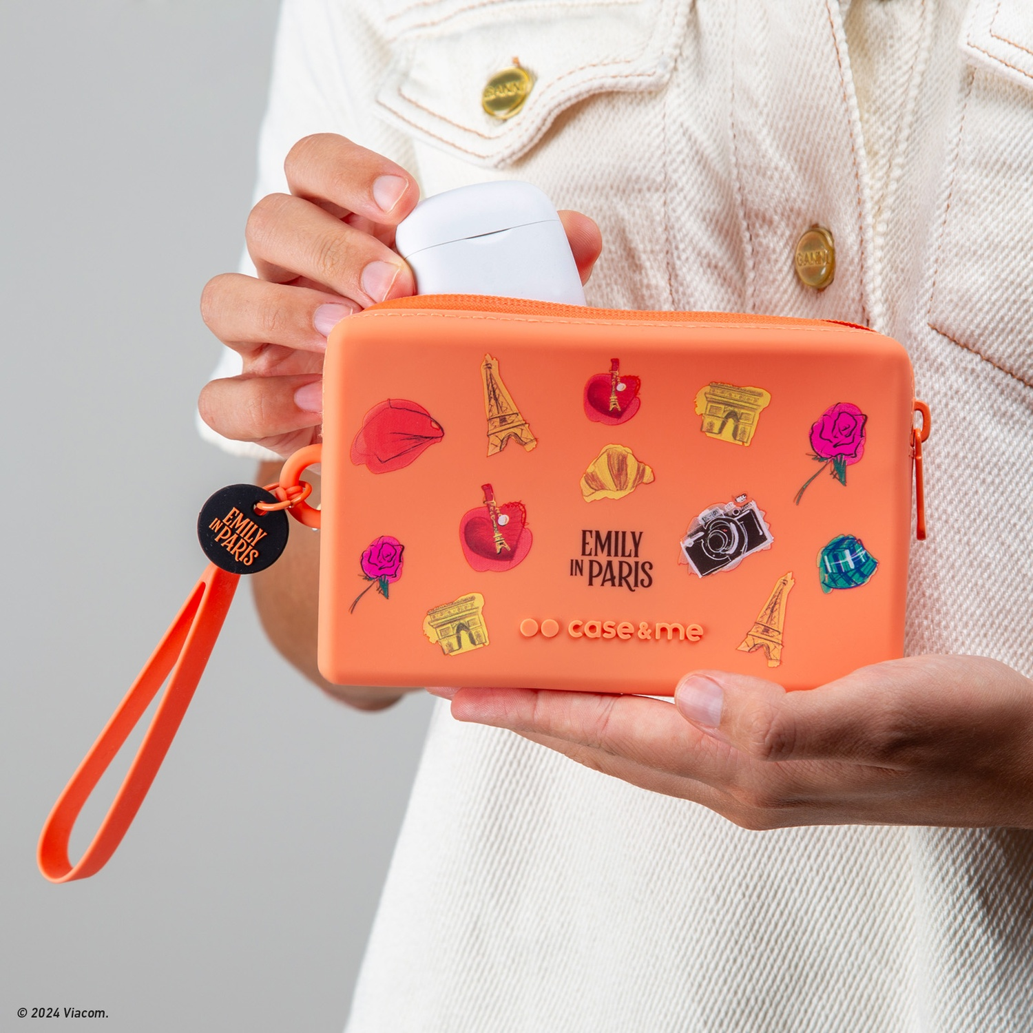 Una persona sostiene una funda naranja con pegatinas de monumentos de París. Dice 'Emily in Paris'. Dentro hay una funda blanca para auriculares.