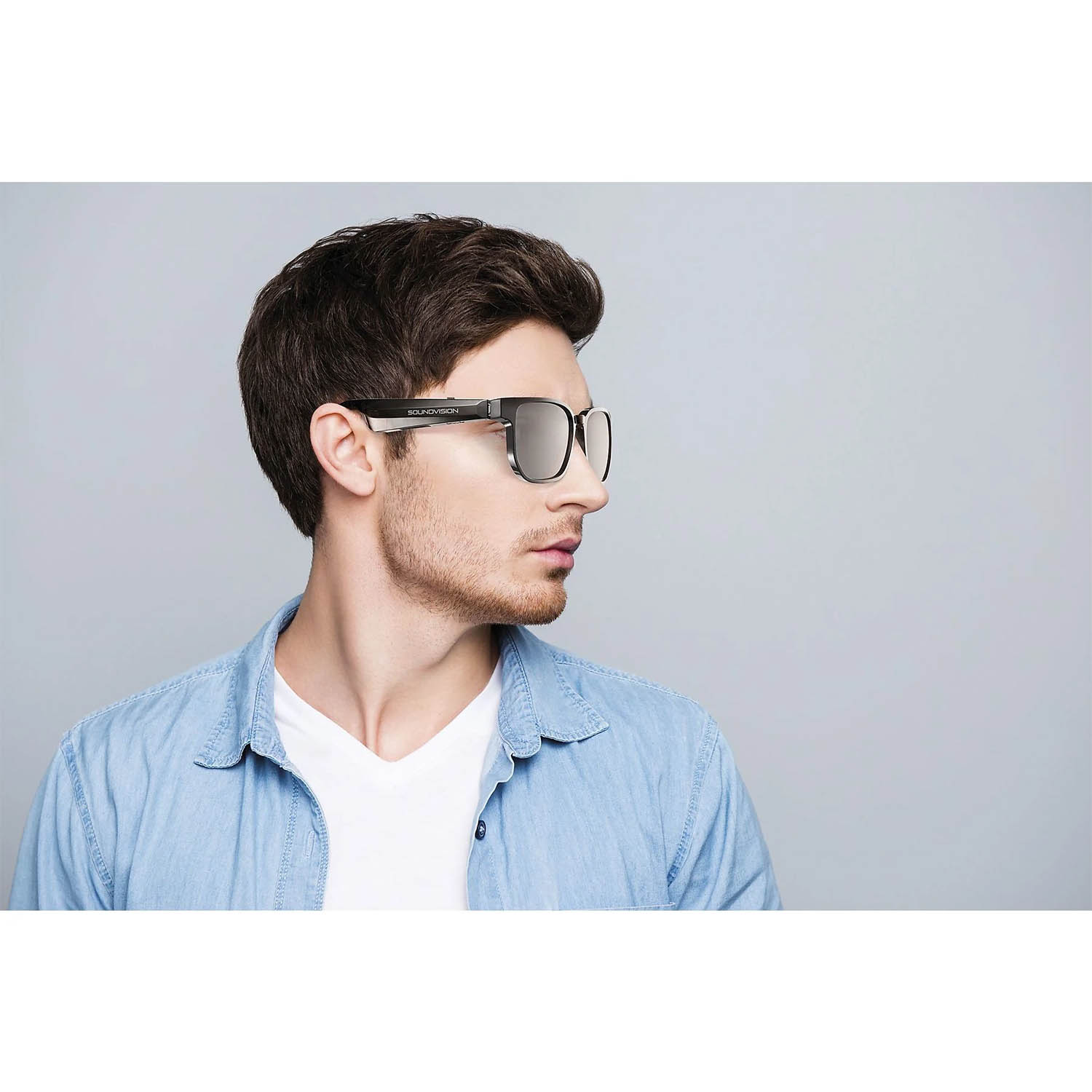 Un uomo con occhiali, camicia di jeans e maglietta bianca. Gli occhiali hanno finitura a specchio e lo sfondo è grigio.
